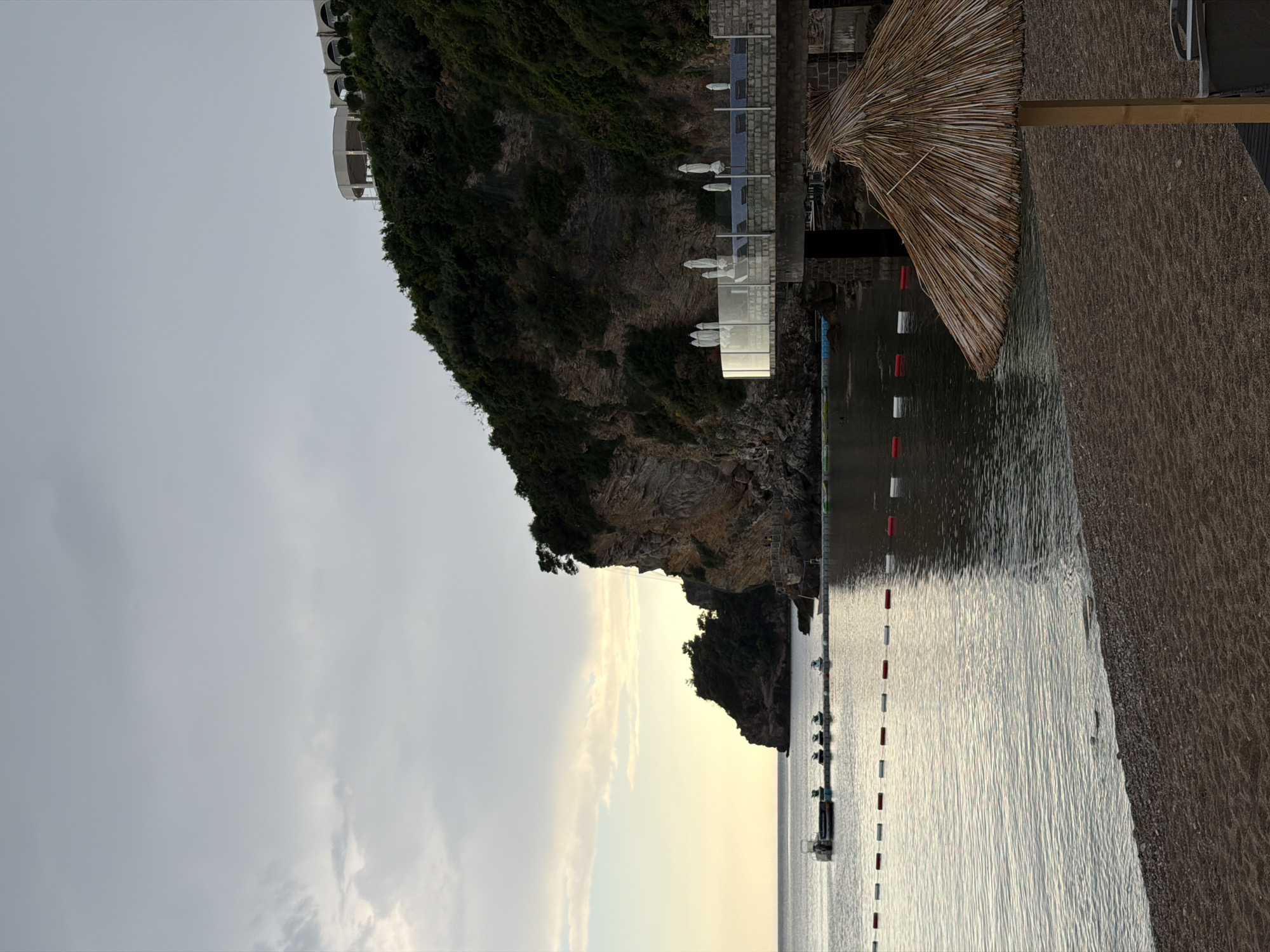 The image captures a beach scene in Budva, Montenegro, featuring a rocky cliff rising from the coastline. The cliff is covered with vegetation and has a building complex situated at its top. A beach runs alongside the water, with a line of red and white buoys marking the swimming area. The water appears calm. A beach umbrella with a thatched roof is also visible. The sky is overcast, suggesting a cloudy day. Several boats are docked near the buoys.