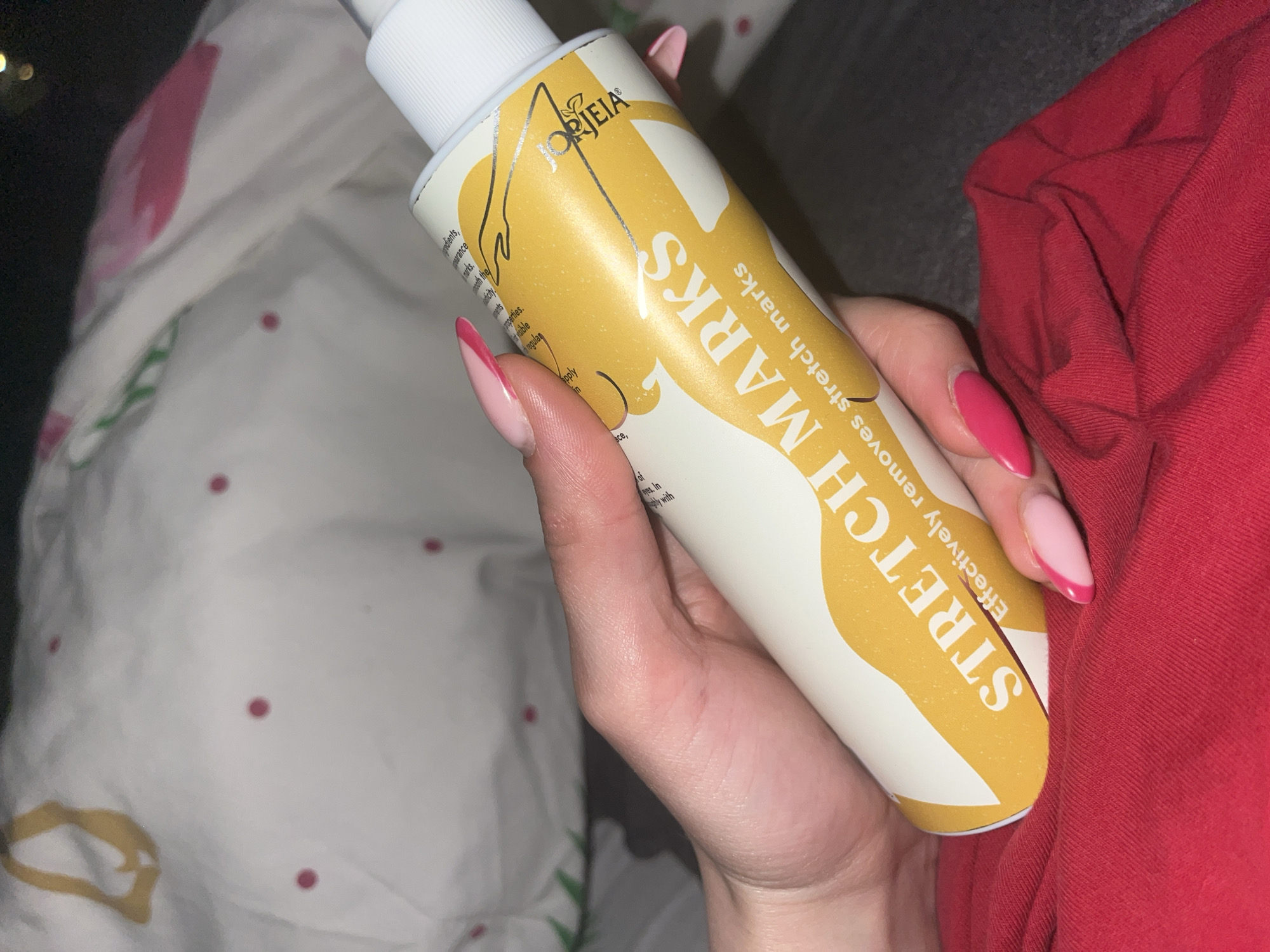 A hand holds a white bottle labeled "JORJEIA STRETCH MARKS Effectively removes stretch marks". The bottle has a gold design. The hand has manicured nails with pink tips. The background includes a patterned bedspread and a red fabric. There is no indication this image was taken in Pernik, Bulgaria.