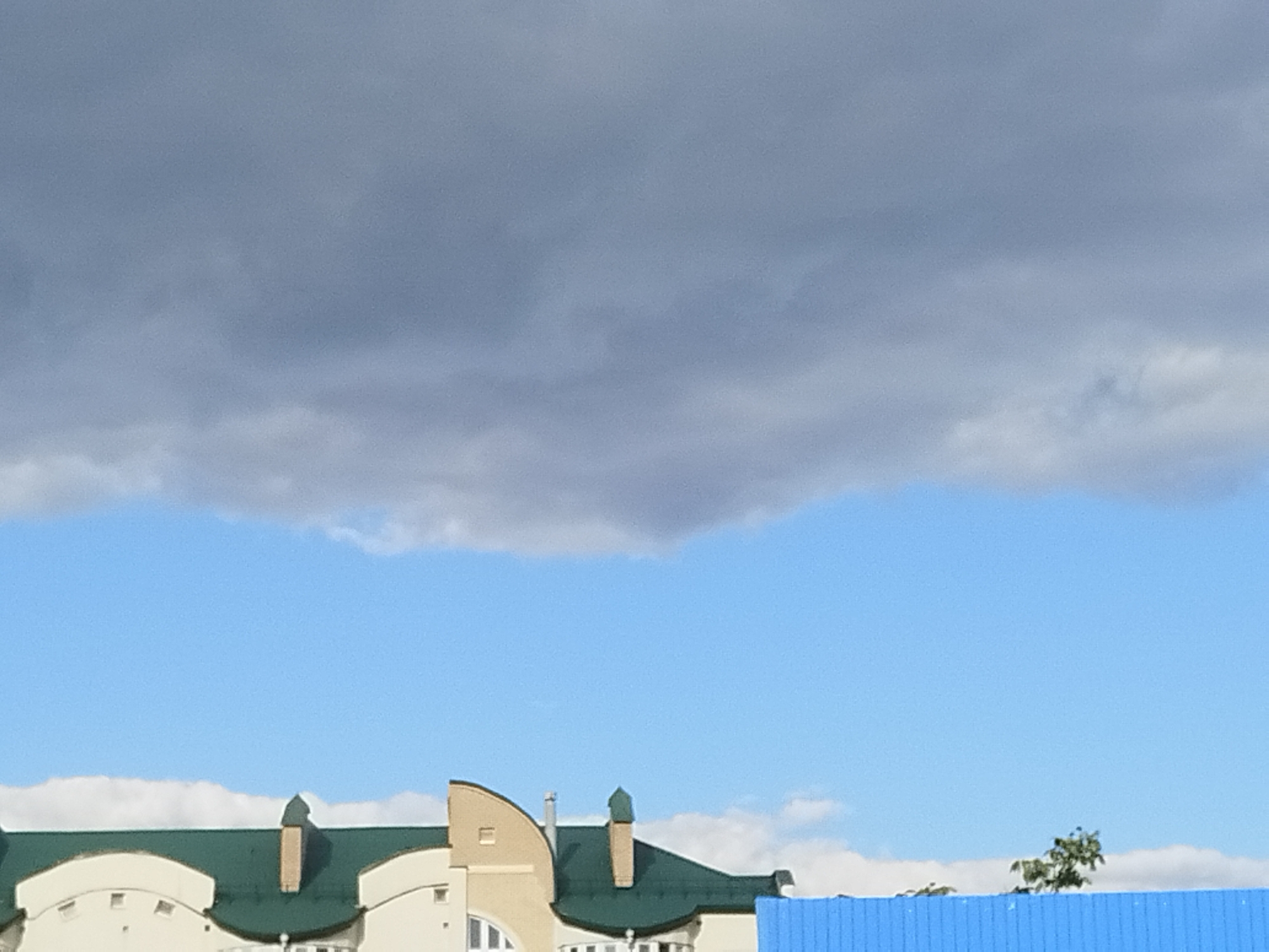 The image captures a section of buildings with green roofs and distinctive architectural features, possibly residential or commercial structures. The sky above is a mix of dark, overcast clouds and lighter blue sky with some wispy white clouds. In the foreground, a bright blue corrugated metal fence is visible, suggesting a boundary or construction area. A small tree with green foliage is seen to the right of the fence. The architecture of the buildings, particularly the pointed roof elements and the curved facade on the left, hints at a European style. Without further information or context, pinpointing the exact location within Brest, Belarus, is not possible from the visual data alone. The scene appears to be an outdoor urban or suburban setting on a day with changing weather conditions.