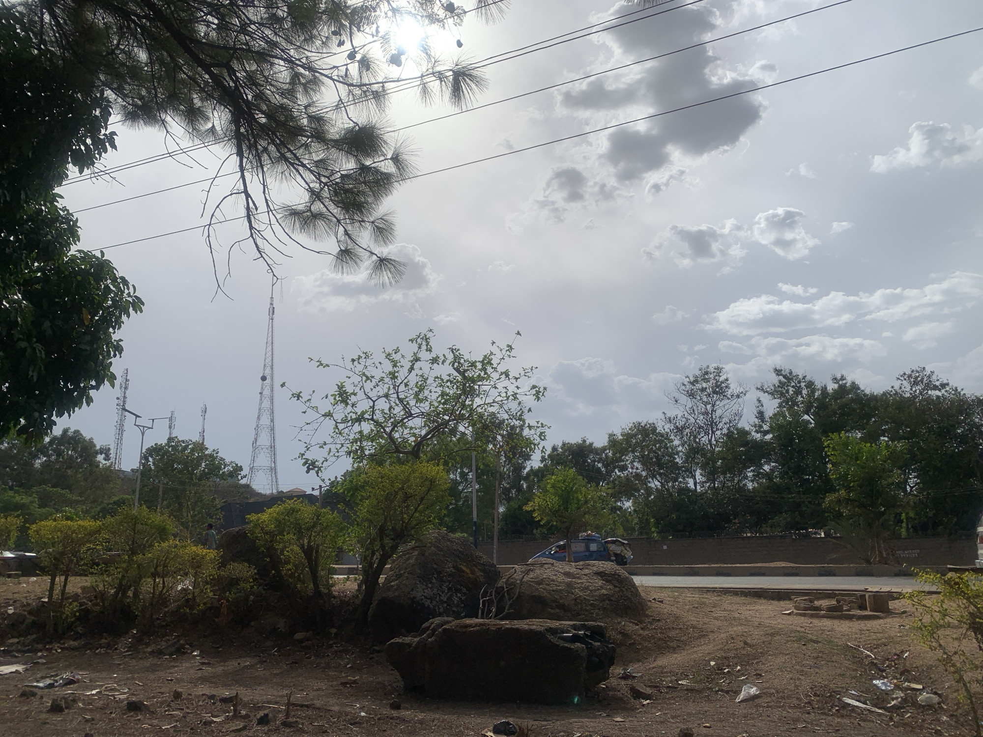 This outdoor image depicts a landscape under a bright, partly cloudy sky. The sun, partially obscured by clouds, shines from the upper left, casting a strong light. Dense foliage from a tree frames the top and left of the image, with needle-like branches extending into the sky. Overhead utility wires traverse the frame.
In the middle ground, several large, dark boulders are prominent, surrounded by smaller green trees and bushes. The ground in the foreground and around the boulders is unpaved, consisting of dirt, scattered rocks, and debris. Beyond the rocks and vegetation, a paved road with a concrete curb and a low wall runs horizontally across the middle of the frame. A blue vehicle, possibly a small bus or van with items strapped to its roof, is visible on the road, alongside a partially visible lighter vehicle on the far right.
On a distant hill, multiple tall communication towers are visible, surrounded by trees. A street light or utility pole also stands near the base of the hill. A lone figure, possibly a child, dressed in light clothing, is standing among the trees and rocks on the left side, near the base of the hill.
The time of day appears to be midday, indicated by the high sun and overall brightness despite the cloud cover. The scene combines natural elements like trees, rocks, and dirt with human infrastructure such as roads, vehicles, and communication towers, suggesting a semi-urban or transitional environment. No legible text is visible.