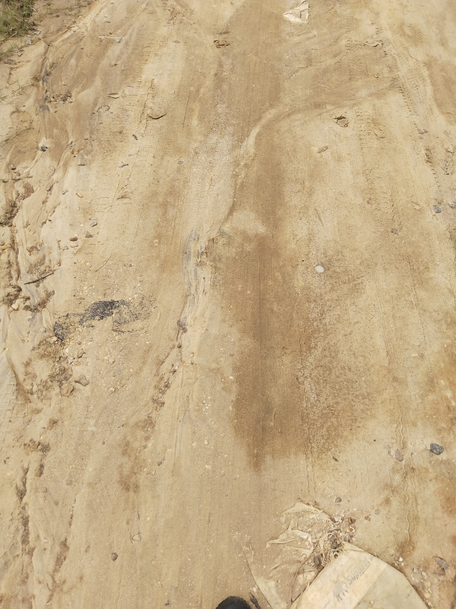 The image shows a section of a dirt road in Uyo, Nigeria. The road surface is primarily composed of sand and loose earth, exhibiting visible tire tracks and areas of erosion. There are small stones and pebbles scattered across the surface. A piece of torn, possibly burlap, fabric lies partially buried near the bottom right corner of the image. There are some dark marks or stains. No people or animals are visible.