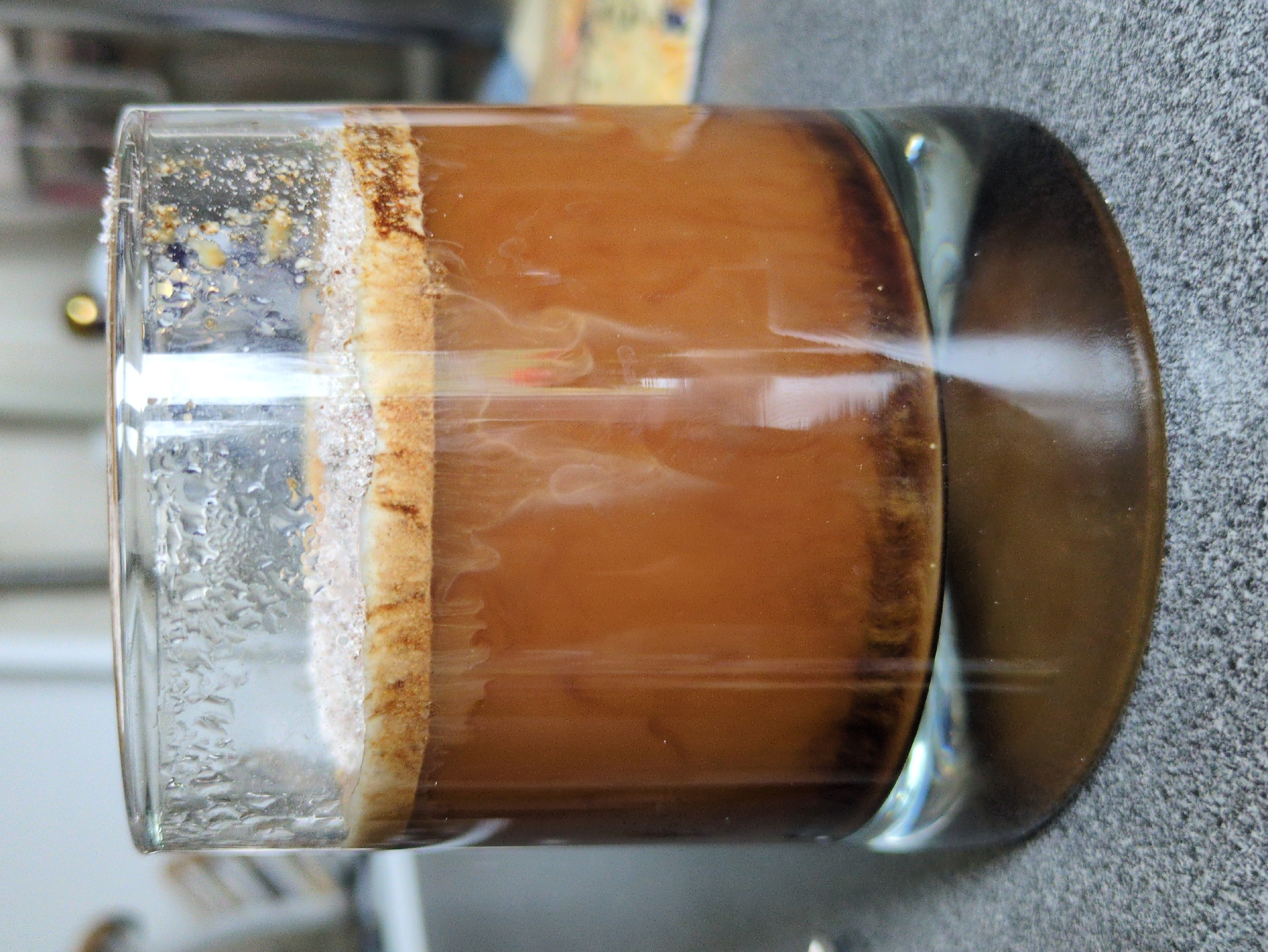 A close-up, eye-level shot shows a clear glass tumbler filled with a dark brown beverage, likely coffee or a coffee-based drink. The drink has a layer of frothy, lighter brown crema on top, sprinkled with what appears to be sugar or fine coffee grounds. Condensation is visible on the outside of the glass, with some larger droplets clustered near the rim. The liquid inside has swirls of lighter and darker brown, indicating movement or mixing. At the bottom of the glass, a sediment of dark brown particles has settled. The tumbler rests on a speckled gray surface, possibly a countertop. The background is blurred and out of focus, with hints of indoor lighting and structures.