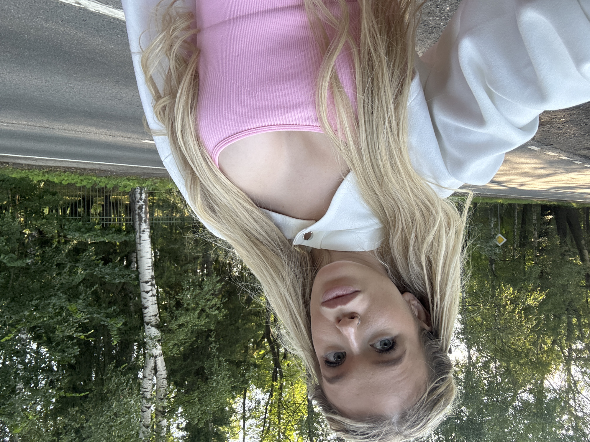 A young woman with long blonde hair, wearing a pink tank top and a white open jacket, poses for a selfie outdoors. She has blue eyes and a nose ring. Behind her, a paved road runs parallel to a tree-lined area with a white birch tree prominently featured. A road sign with a yellow diamond shape is visible in the background. The environment suggests a suburban or semi-rural setting, possibly on the outskirts of Pavlovsk, Russia, given the presence of mixed deciduous and coniferous trees and the general atmosphere. The lighting indicates it is daytime, with sunlight filtering through the trees.
