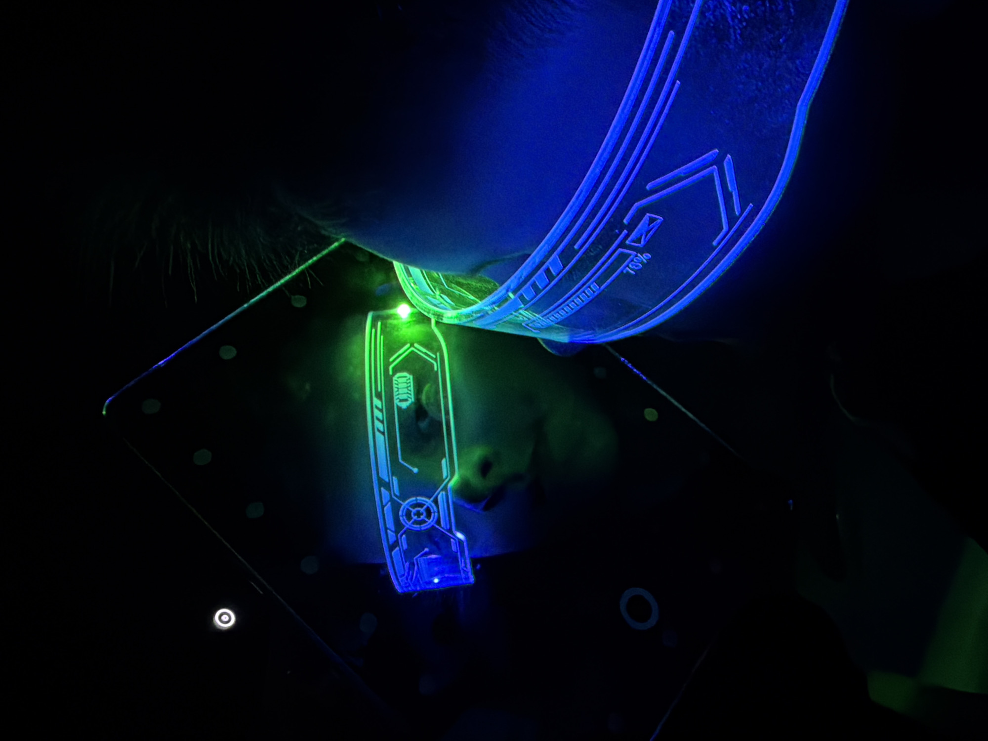 A close-up shot in a dark environment features a child's face illuminated by two glowing, transparent objects with futuristic designs. The upper object, curved and blue, rests against the child's forehead. The lower object, a flat rectangle displaying a green and blue design, is held near the child's nose. Small white circles are visible on the rectangle. The child's nose and mouth are visible, and they appear to be looking at the illuminated objects. Sparse hair is visible near the top edge of the rectangle. The location is not discernible from the visual information provided.