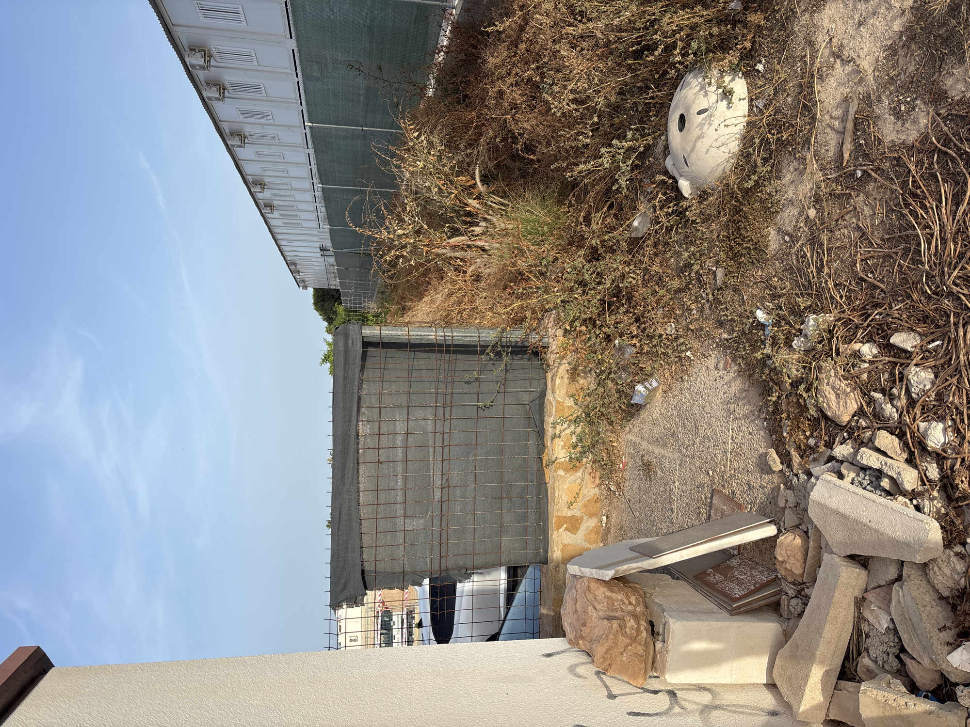 The image depicts an outdoor scene in Orihuela, Spain. In the foreground, a pile of construction debris including concrete blocks and rocks is visible near the base of a textured beige wall on the left. Behind the debris, a partially covered structure with wire mesh and dark netting stands on a stone foundation. To the right of this structure, overgrown, dry vegetation dominates the scene, with some litter scattered amongst the plants, including a discarded white ceramic object resembling a toilet bowl. In the background, a long, white prefabricated building with multiple windows and air conditioning units runs parallel to a green chain-link fence. The sky above is clear and blue with a few wispy clouds. A white car is partially visible behind the fenced structure.