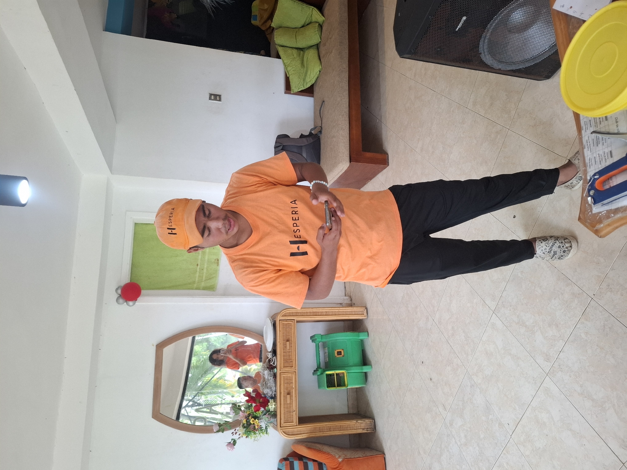 A young male, wearing an orange "Hesperia" cap and an orange t-shirt with the same logo, stands in a room and looks at his phone. He is wearing black pants and patterned shoes. Behind him, a mirror reflects two other individuals in orange shirts. In the background, there is a couch with green pillows, a rattan console table with flowers and a green blower, and a large speaker. The room has tiled floors and white walls. A red and silver balloon decoration is visible on the wall. The scene appears to be indoors in a casual setting, possibly at a resort or event venue in Playa El Agua, Venezuela.
