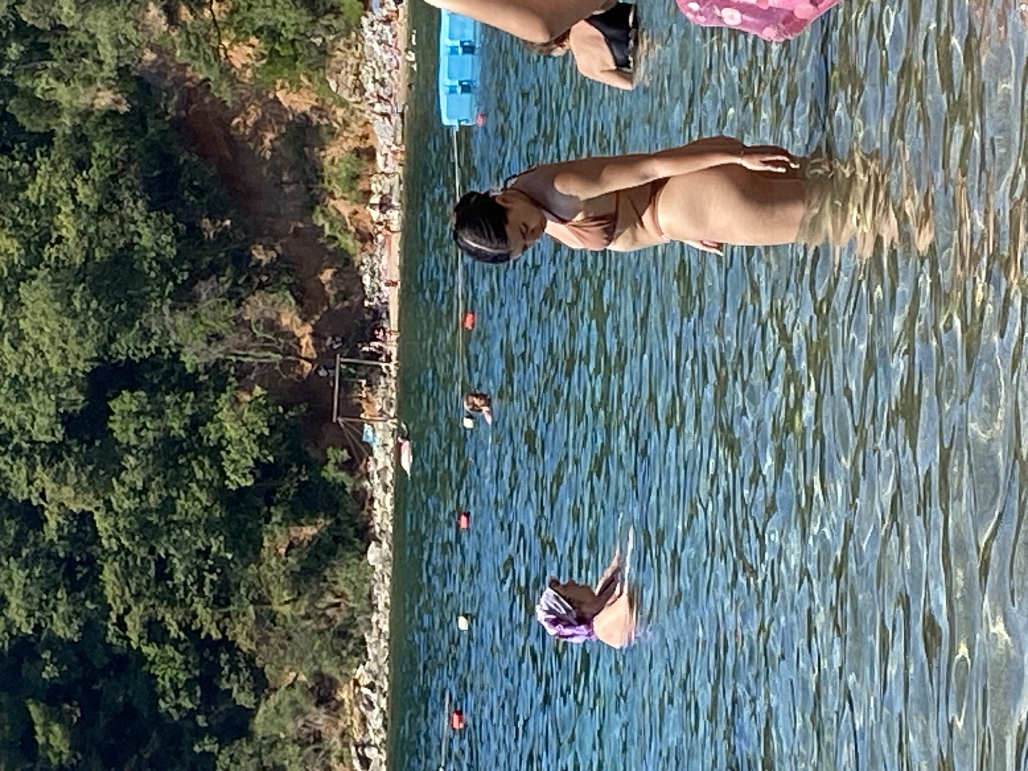 The image depicts a sunny day at a beach, likely in Budva, Montenegro, given the hilly, tree-covered coastline and the clear blue water. In the foreground, a young woman wearing a brown bikini stands in the shallow water, facing away from the camera, with her left hand resting on her hip. To her left and slightly further back, an older woman in a purple headscarf and a swimsuit is also in the water, looking to her right. Further out in the water, other individuals can be seen, including a person in a black swimsuit. The background shows a rocky shoreline with dense green trees and some scattered buildings or structures on the hill. There are buoys in the water, marking off an area, and a large blue floating structure is visible on the right side of the image. The water is calm with visible ripples and light reflections. The overall scene conveys a relaxed atmosphere of a summer day at the seaside.