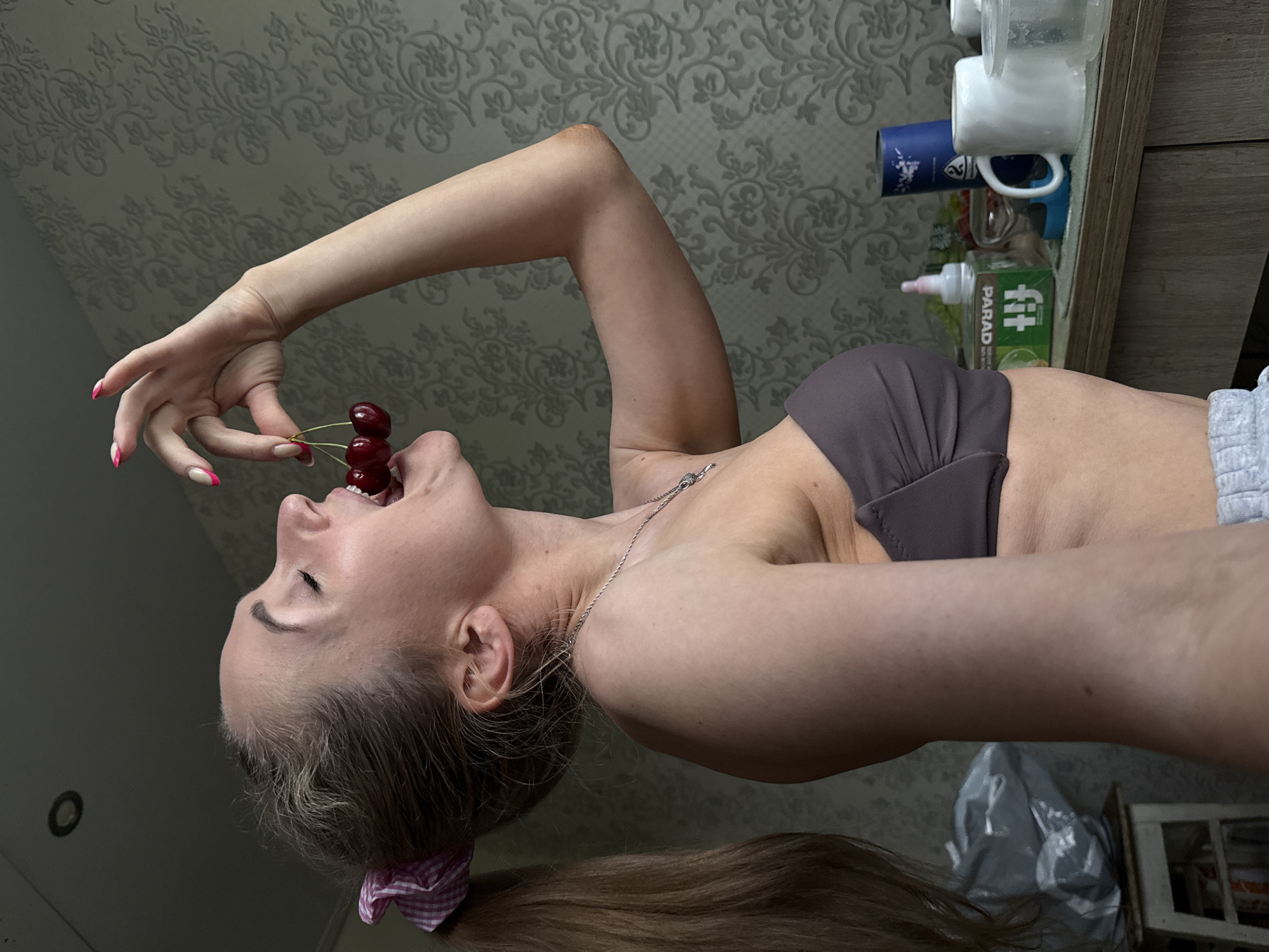 A light-skinned woman is holding a cluster of cherries to her open mouth in what appears to be an indoor setting. The woman is wearing a gray-brown sports bra and gray shorts, and has a ponytail secured with a pink and white checkered hair tie. She is wearing a silver chain around her neck. The nails on her left hand, which is holding the cherries, are painted with a French manicure style, featuring a pink tip.
Behind the woman is a wall with a light gray patterned wallpaper. To the right of the woman are various items including white mugs, a blue mug, and a box with the word "fit" displayed prominently. Further to the right is a light-colored wooden cabinet.
There is no contextual information suggesting the location is in the city of Chaikovsky, Russia.