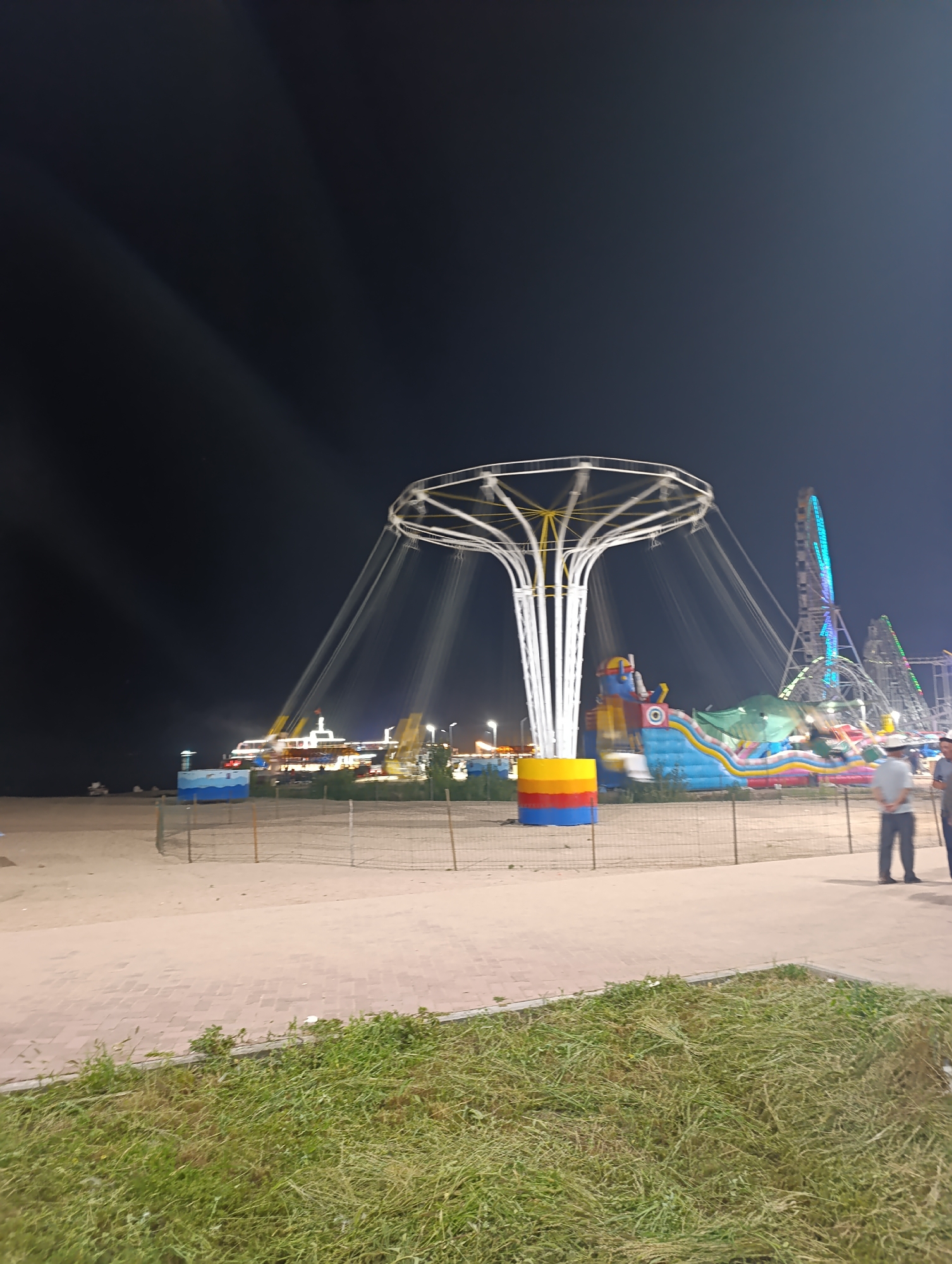 An amusement park is depicted at night on a sandy beach in Bosteri, Kyrgyzstan. A swing ride, a Ferris wheel, and a roller coaster are visible. The swing ride is in the foreground, with its white poles and yellow center illuminated. People are standing on a paved walkway in the lower right corner of the image, observing the amusement park. The beach is empty except for the amusement park attractions. The sky is dark and clear, with some light streaks visible, possibly from the lights of the rides or a long exposure effect.