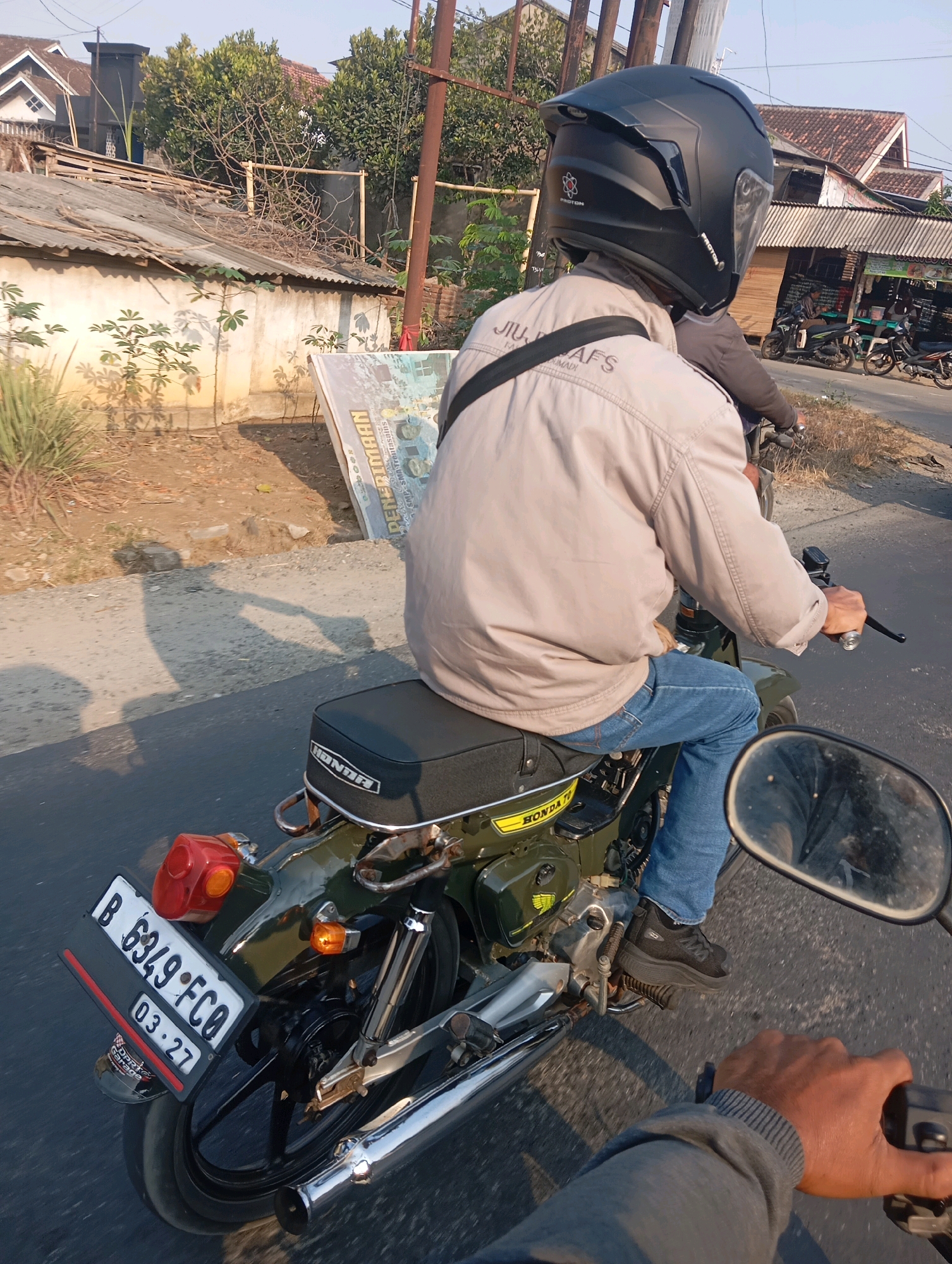 The image shows a perspective from the back of a motorcycle in motion, looking forward. The driver, wearing a black helmet and a light-colored jacket over blue jeans, is seated on a vintage-style green Honda motorcycle. The motorcycle's license plate reads "B 6349 FCO" with "03-27" below it. A prominent "HONDA" logo is visible on the motorcycle's seat. The background reveals a rural or semi-urban environment with houses, trees, and a dirt road. In the far distance, other motorcycles are parked outside a building that appears to be a shop or market. The sunlight suggests it is daytime. The scene takes place on a paved road, likely in the outskirts of Ganjar Asri, Indonesia, given the context.