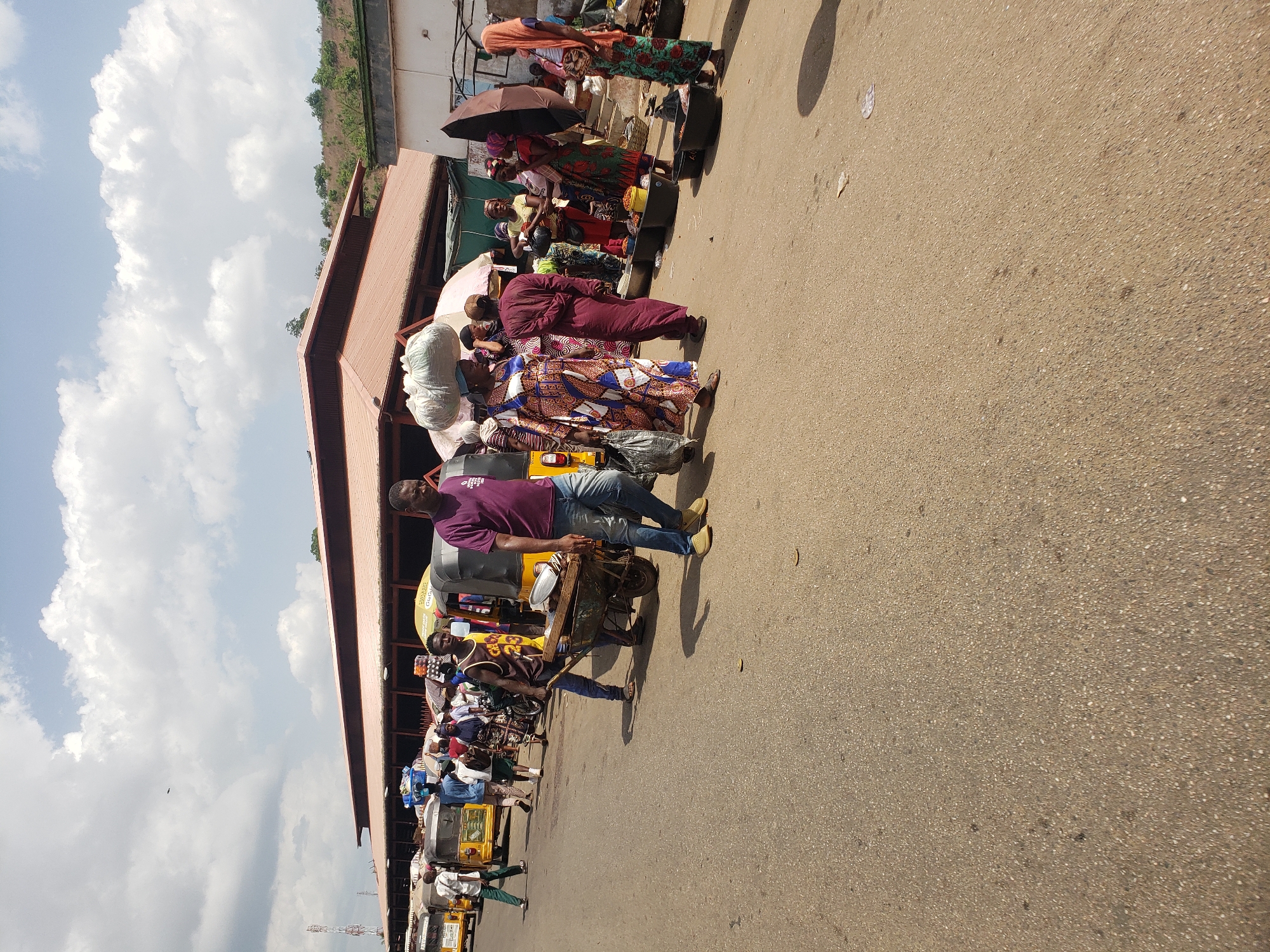 The image depicts a busy street scene in Lokoja, Nigeria. A row of people, primarily women in colorful African prints, stand adjacent to a building with a red roof, possibly a market. Some individuals have goods for sale, including items in large sacks and produce in containers. A man in a purple shirt and jeans is pushing a heavily laden cart. Several auto-rickshaws (tuk-tuks) are parked along the street. The sky is partly cloudy. The street is paved, with some debris and indistinct objects on the surface.