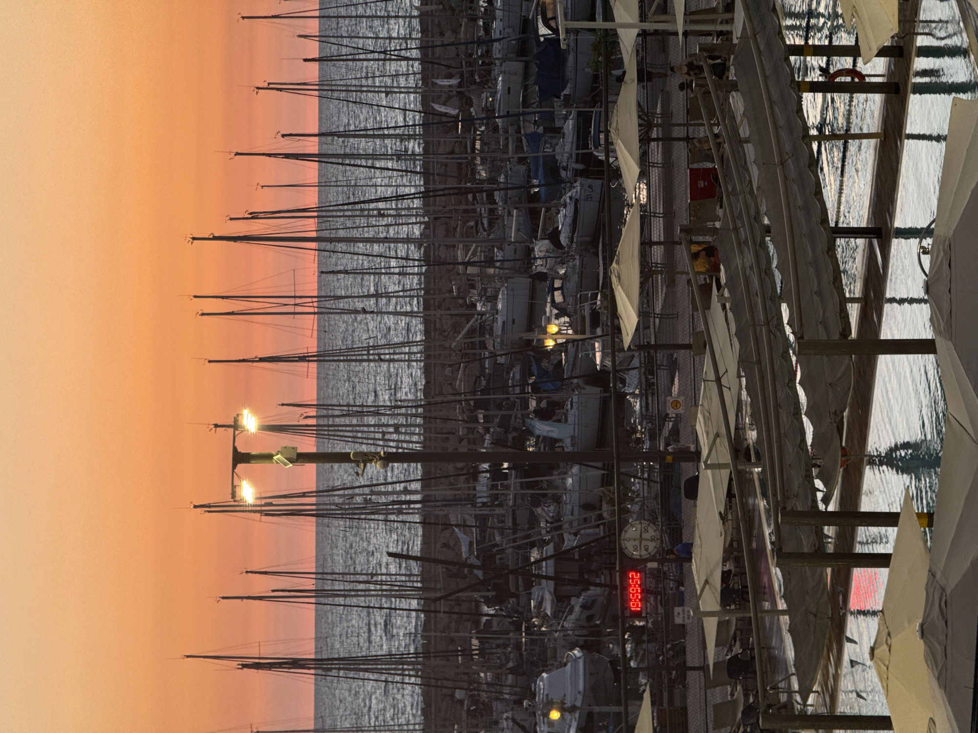 The image captures a harbor scene at sunset in Tel Aviv, Israel. The sky is a vibrant orange, transitioning to a softer yellow near the horizon. Silhouetted against this colorful sky are numerous sailboat masts, suggesting a marina. In the foreground, a swimming pool area is visible with lounge chairs and umbrellas, indicating a resort or hotel. A digital clock on a pole displays "19:59:52," and a round gauge with needles points to approximate positions. Several boats are docked in the harbor, some with lights on. The overall atmosphere is serene, with the gentle glow of the sunset reflecting on the water.