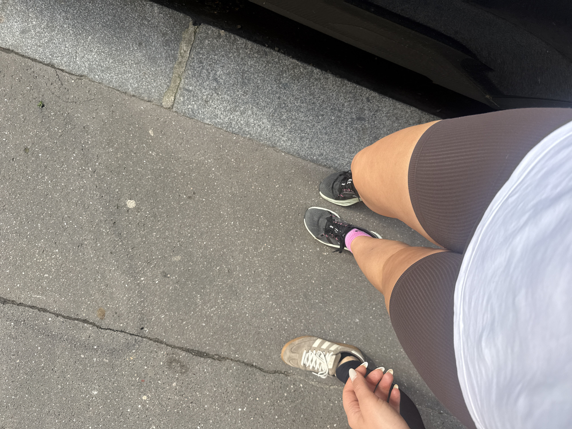 The photograph, taken at a downward angle, presents a partial view of a person standing on a city street. The individual is wearing brown ribbed shorts and a white shirt, which is mostly out of frame. Their feet are clad in two different types of athletic shoes: one is an Adidas sneaker with a light gray upper and white stripes, paired with a black sock, and the other is a dark gray running shoe with pink detailing. The person is holding a thin black object, possibly a phone cord or similar item, in their hand.
The ground is paved with gray asphalt, showing signs of wear such as cracks. A curb made of granite blocks is visible along the top edge of the frame, adjacent to a dark surface that is likely a vehicle. The scene conveys a sense of urban everyday life.
There is not enough information in the image to determine if it was taken in Paris, France.