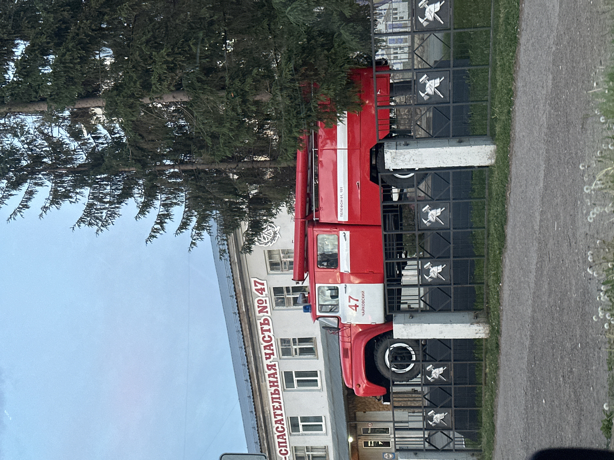 A red fire truck with the number "47" is parked in front of a building that reads "SPASATELNAYA CHAST' NO 47" (Rescue Unit No. 47) in Russian. The truck also has the word "CHAIKOVSKY" written on it, indicating the city where it is located. A pine tree is visible in the foreground. A metal fence separates the fire truck from the sidewalk.