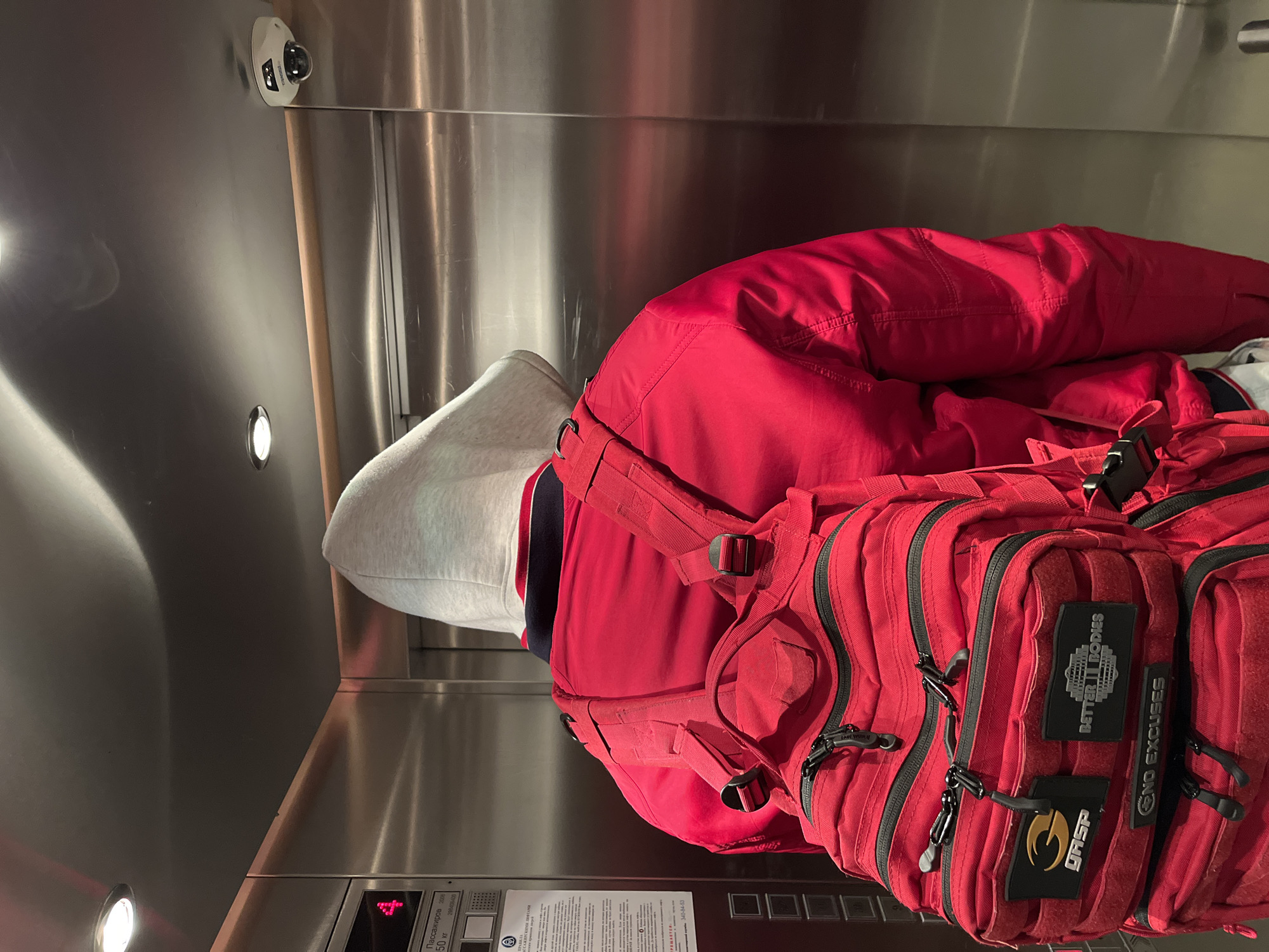 A person wearing a grey hoodie and a red jacket is standing in a metal elevator. The person is also wearing a red backpack with black zippers and patches, including "BETTER BODIES", "GASP" and "NO EXCUSES". A security camera is mounted in the upper portion of the elevator. An elevator control panel with an illuminated "4" is visible. Text in Russian is located on the elevator wall. Based on the Russian text, the elevator has a capacity of 50 kg and was installed in 2008. The phone number 340-84-53 is also visible on the wall. It is not possible to identify the location in Saint Petersburg, Russia, without additional information.