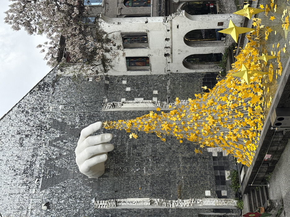 The scene depicts an outdoor art installation located in Yuzhong District, China. A large, pale grey hand sculpture extends from the upper section of a dark, textured building facade on the left. This facade features a fragmented, distressed appearance with exposed brickwork and a subtle "YW" mark. The hand's fingers appear to be interacting with the apex of a large, conical structure densely covered in numerous yellow five-pointed stars attached by thin rods or wires. This star sculpture occupies a ground-level planter area. Scattered around its base and extending towards the foreground are additional yellow stars and several larger, freestanding yellow star-shaped sculptures. To the right, an older, light-colored building with multiple arched windows and ground-level entrances, some containing stained glass-like panels, is partially visible. A mature tree with light-colored blossoms partially obscures the upper portion of this building. A stone wall separates the immediate foreground, which includes a dark waste receptacle, from the main installation. Steps descend on the far left. The sky is uniformly overcast. No individuals are present in the frame.