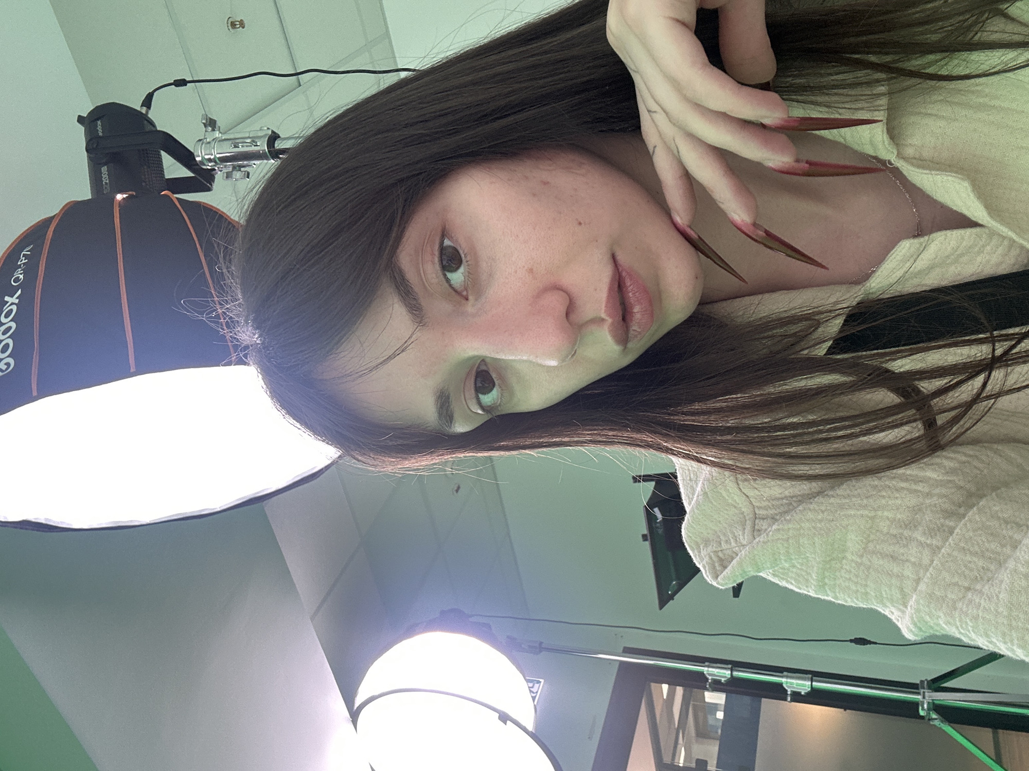 A woman with long, dark hair and long, pointed fingernails is posing in a studio setting, likely in Belgrade, Serbia. She is wearing a light-colored top. There are two studio lights visible in the frame, one with a softbox and another with a different lighting apparatus. The ceiling and walls of the studio are visible, as is some lighting equipment mounted on a rail.