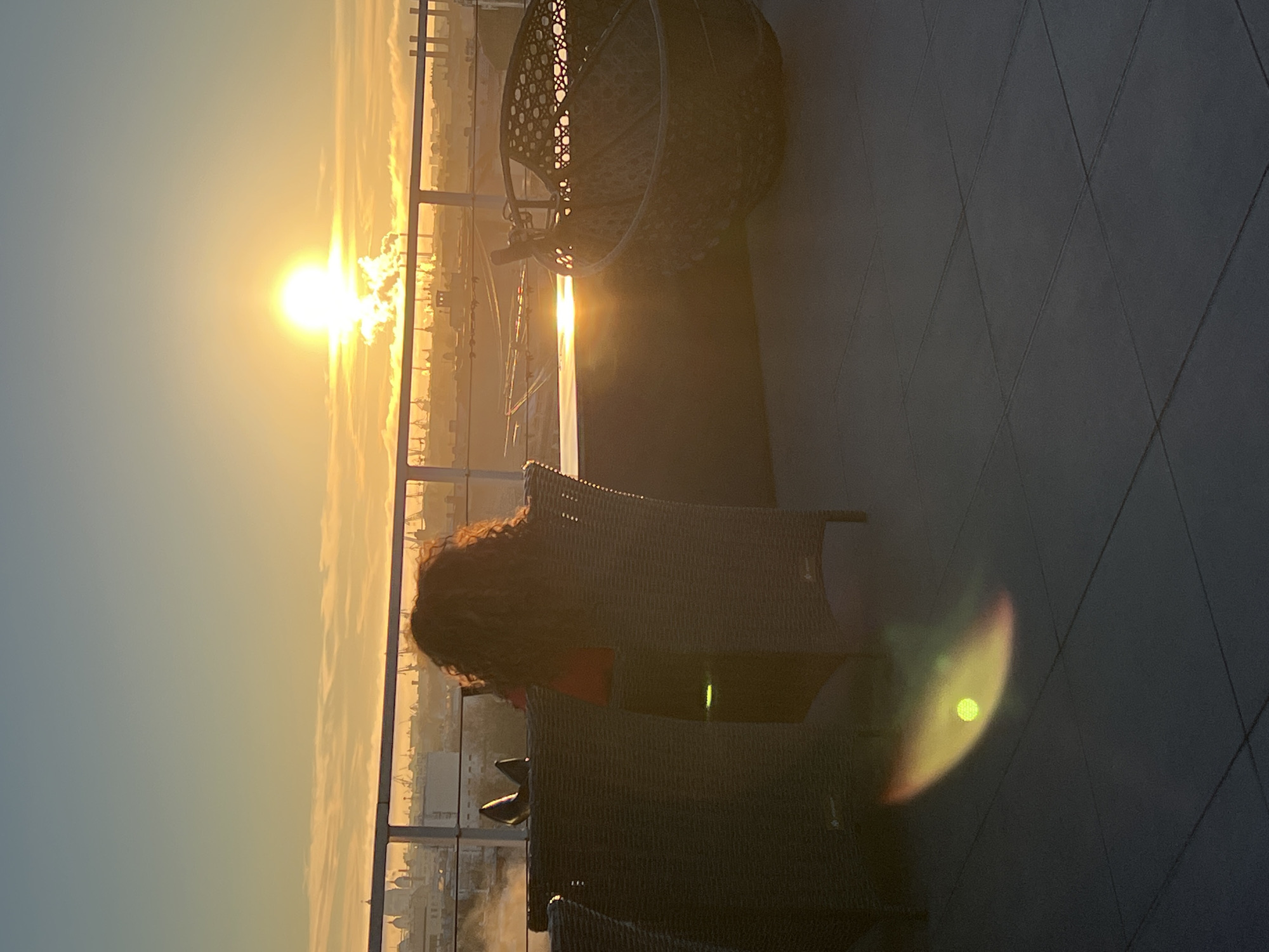 A person with curly hair sits in a wicker chair on a balcony overlooking the city of Saint Petersburg, Russia. The sun is setting over the city, casting a golden glow. A hanging chair is in the background. The balcony has a dark tiled floor.