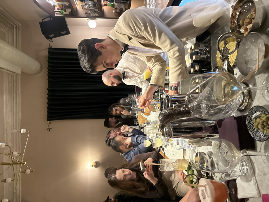 The image depicts the interior of Dumbarton, a cocktail bar located on Saryan Street in the Kentron district of Yerevan, Armenia. The scene is dimly lit, characterized by light beige walls, dark green velvet curtains on the left, and a gold-toned multi-bulb chandelier overhead.
Approximately ten adult individuals are visible. Two male bartenders are positioned behind a light-colored bar counter. The primary bartender, right-of-center, wears a beige blazer, a dark bow tie, and a digital watch on his left wrist. He is actively engaged in mixing a drink, holding a small silver bar tool over a glass while also manipulating a bottle containing a yellow liquid. A second male bartender, with a shaved head and wearing a white shirt with a dark bow tie, is partially visible behind the primary bartender, observing the process.
The bar counter in the foreground is populated with various objects: multiple cocktail glasses (some containing drinks such as a reddish-orange concoction with foam and ice, and clear beverages with ice), several metallic cocktail shakers (one featuring a distinctive dark, ornate bird-head handle), a bowl of green olives/pistachios, a bowl of mixed nuts, and bowls of ice. Small bottles of spirits and mixers are arranged on shelves behind the bar.
On the left side of the frame, a female patron with long brown hair, dressed in a brown top and beige skirt, is seated at the bar, focused on her smartphone. A tall glass with a straw stands on the counter in front of her. Next to her, a male patron with dark hair and a beard, wearing a blue shirt, leans forward, appearing to observe or engage with other patrons. Further back, additional patrons are partially visible seated or standing against the dark green curtains, engaged in social interaction. The overall environment suggests an upscale and intimate bar setting.