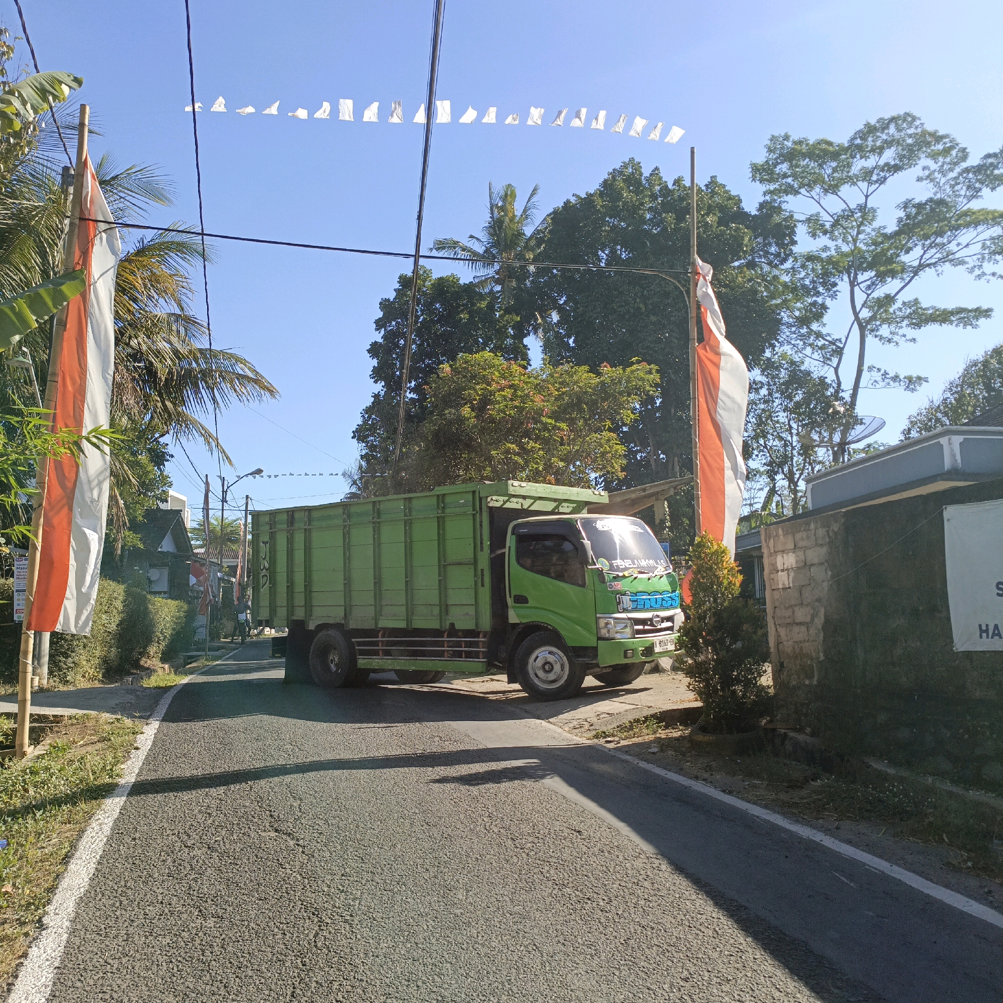 The image depicts a scene on a paved road in Salatiga, Indonesia. A green truck is parked on the right side of the road, facing left. The truck has an open cargo bed. In the foreground, the road is visible with a white line marking the edge. To the left of the road, there is a ditch and some vegetation. Two tall poles with Indonesian flags (red and white) are planted on the left side of the road, and a string of small white flags is strung across overhead. In the background, there are houses, trees, and a clear blue sky. The lighting suggests it is daytime, with sunlight casting shadows.