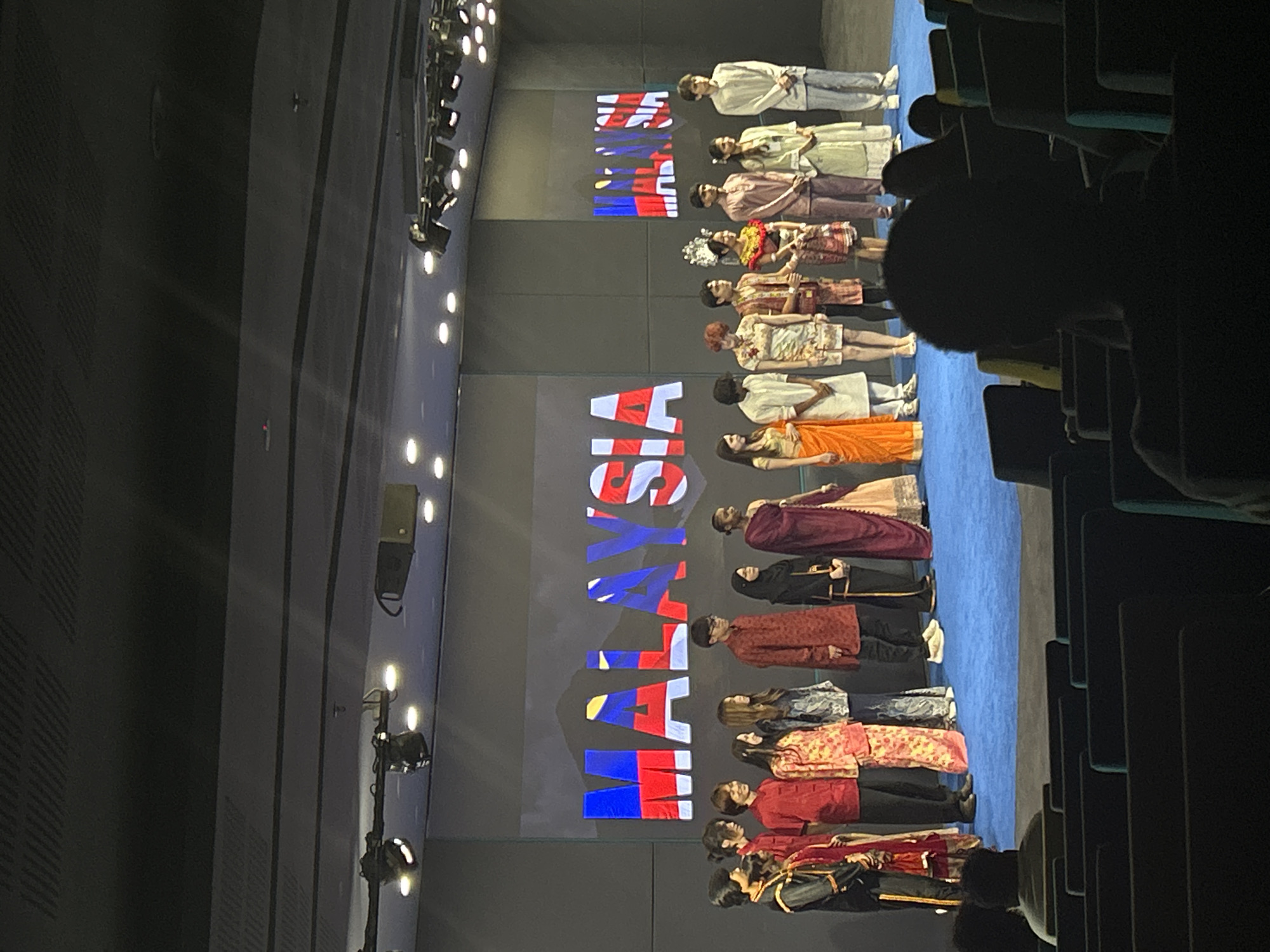 The image depicts a group of individuals standing on a blue-carpeted stage in what appears to be an auditorium or large hall. The backdrop features a screen displaying the word "MALAYSIA" in a stylized font, incorporating the colors of the Malaysian flag. The individuals are dressed in what seems to be traditional Malaysian clothing. Rows of audience seating can be seen on the right, indicating an event or performance taking place. The hall appears to be equipped with stage lighting, visible in the upper left corner. The city of Kuala Lumpur, Malaysia is listed as the location.