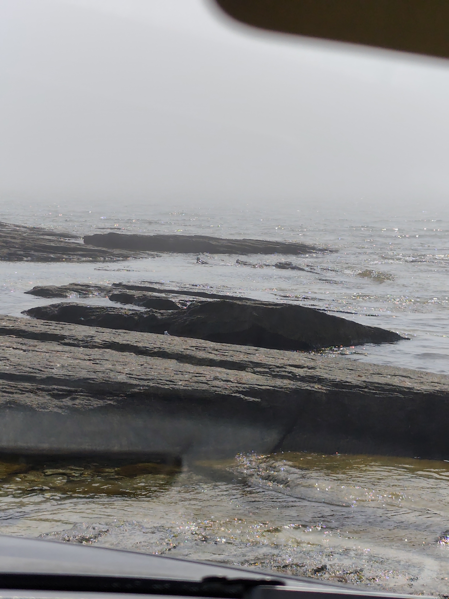 The image shows a rocky shoreline with the Caspian Sea in the background. The water is choppy and reflects the sunlight, creating a sparkling effect. Several large, flat rocks are submerged in the water, with others visible on the shore. The sky is hazy and overcast, with a faint fog obscuring the horizon. There is no indication of human presence or activity in the image. The location is not identifiable as Saiyn Şapağatov, Kazakhstan, as there are no specific landmarks or features that would suggest this location.