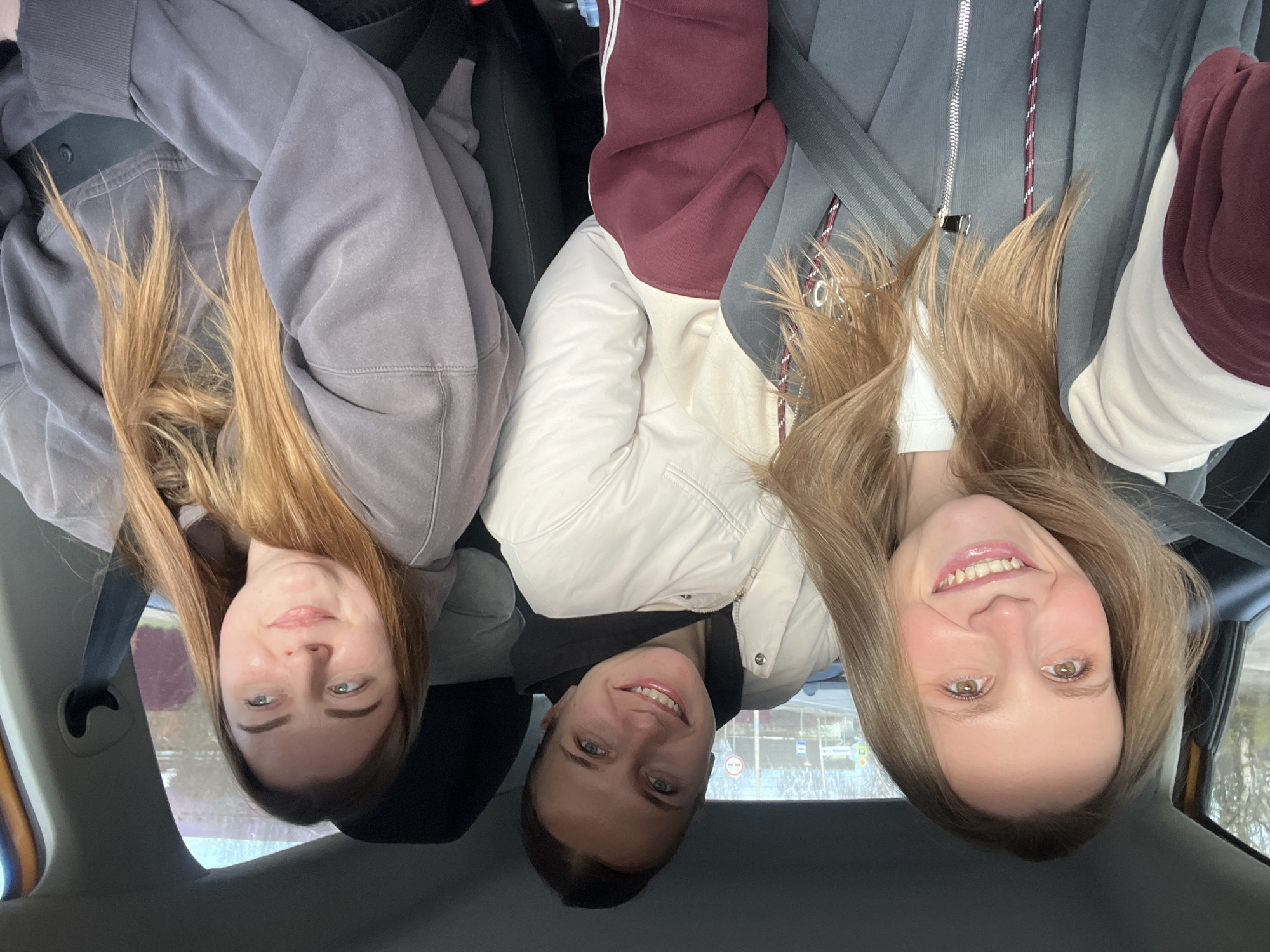 Three young women are captured in a cheerful selfie taken inside a car, likely in Ширяево, Russia. The image, originally oriented upside down, shows them from a low angle, suggesting the photo was taken by one of them looking upwards or from the back seat. All three are smiling, conveying a sense of happiness and camaraderie.
On the left side of the corrected orientation, a woman with long, light brown hair wears a muted grey or purple hoodie. She has light-colored eyes and a gentle smile. In the center, another woman with darker hair, possibly brown, smiles broadly, showing her teeth. She is wearing a black hoodie under a white puffer jacket, and her eyes are also light-colored. On the right, the third woman, with long, light brown hair, smiles widely, revealing her teeth. She is dressed in a grey zip-up jacket with maroon and white sections, and a white shirt underneath. Her eyes appear green or hazel. Seatbelts are visible across the chests of the women on the left and right, indicating they are passengers.
The setting is clearly the interior of a vehicle, with parts of the car's roof lining and seats visible. Through the car windows in the background, a blurred outdoor scene suggests daytime travel, possibly with some urban or natural elements visible but indistinct. The lighting appears natural and bright, indicating it is daytime. No discernible text is clearly visible on clothing or in the background.