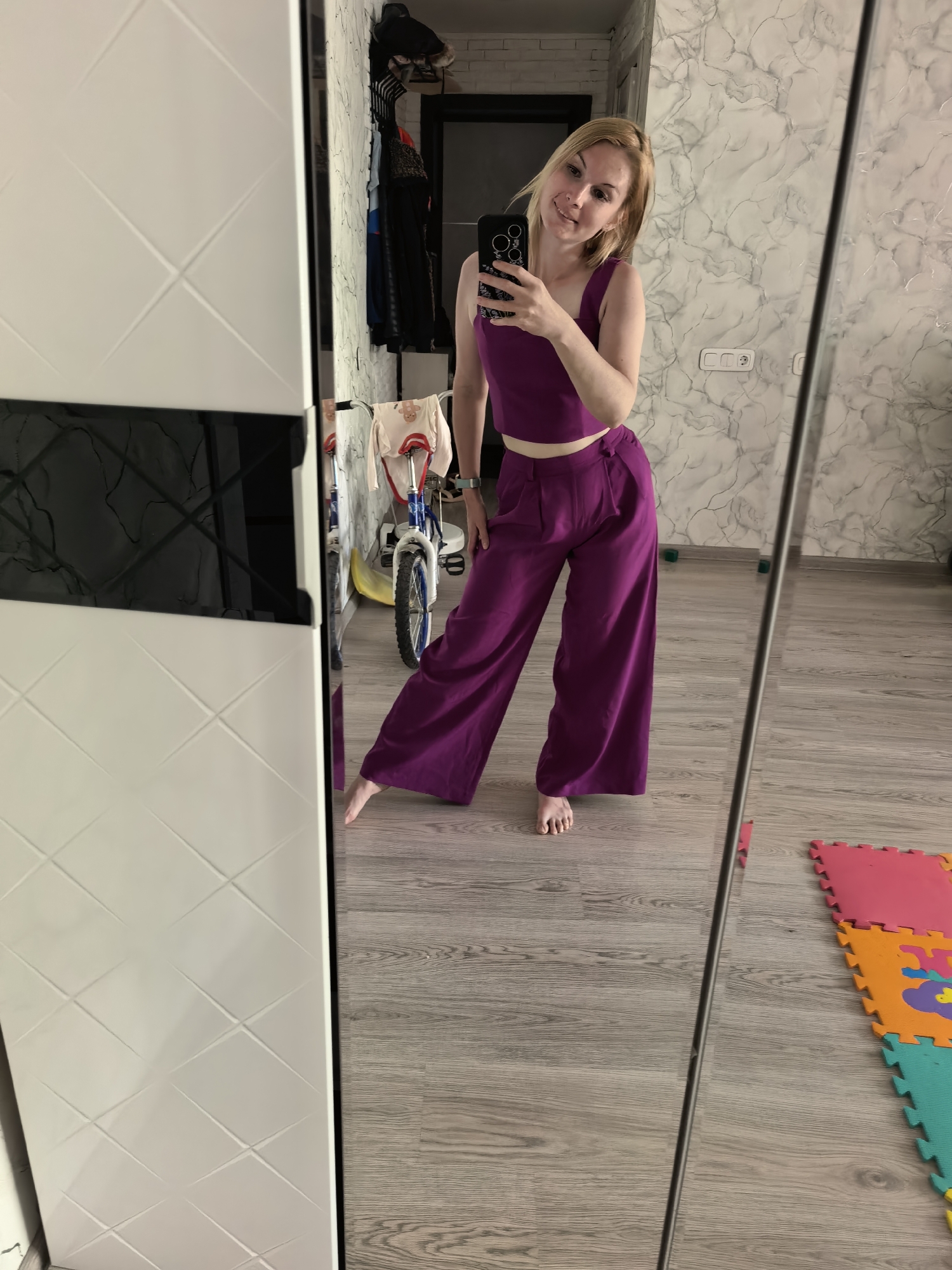 A person with blonde hair is taking a selfie in a mirror. The person is wearing a purple two-piece outfit, consisting of a crop top and wide-legged pants. A cell phone is visible in their hand. They are positioned indoors, likely in a hallway or room of a house. A white wardrobe is visible to the left of the mirror. Behind the person, in the reflection, is a child's bicycle. There is also a wall with a marble-like pattern. In the lower right corner of the reflection, there are colorful foam play mats on the floor. There is no information to determine the location in Sterlitamak, Russia.