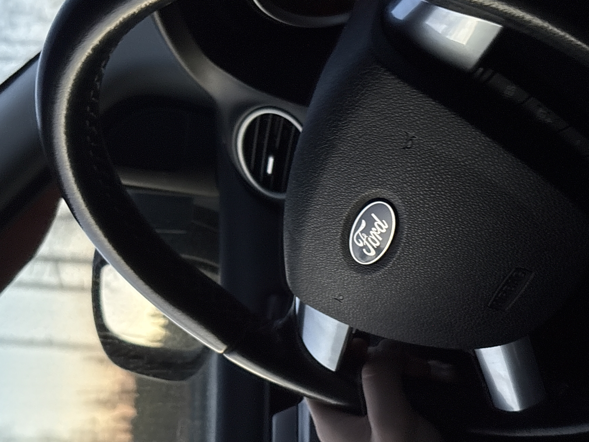 A close-up image shows the steering wheel of a Ford vehicle. The Ford logo is visible in the center of the steering wheel. A ventilation vent is visible behind the steering wheel. The image does not provide any information about the location of Yoksolovo, Russia, or any people or actions.