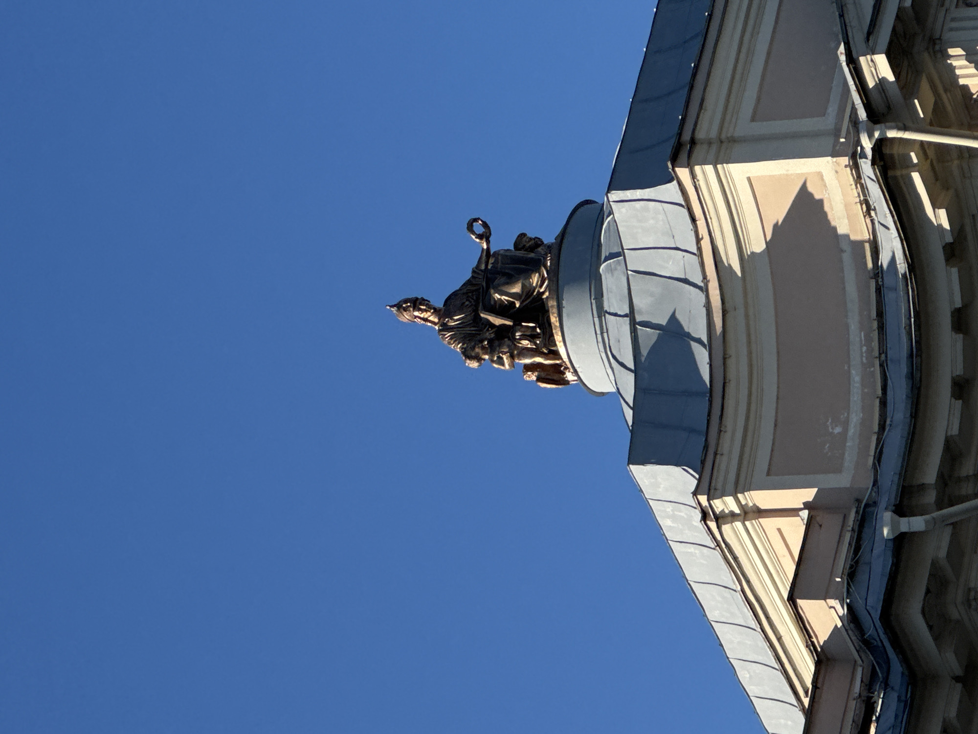 An upward-angled shot captures a bronze or dark metal sculpture adorning a classical or neoclassical building in Saint Petersburg, Russia, against a vibrant, cloudless blue sky. The image is oriented vertically, showing the sculpture on the upper portion of a building, likely a corner or a prominent architectural feature.
The sculpture depicts a figure, possibly allegorical, with draped clothing, holding a laurel wreath aloft with one hand. The figure appears to be seated or kneeling on an ornate base that is part of the building's facade. The details of the sculpture are visible due to strong, direct sunlight, which also creates sharp shadows, indicating a bright day.
The building itself features ornate architectural details, including cornices, moldings, and pilaster-like elements in light tones, possibly stone or stucco. Part of a dark, sloping roof or dome is also visible, with its surface showing individual panels or tiles. The building's structure is grand and historical. No specific activity is occurring as the image focuses on static architecture and sculpture. There is no visible text. The bright lighting and clear sky suggest it is daytime with fair weather.