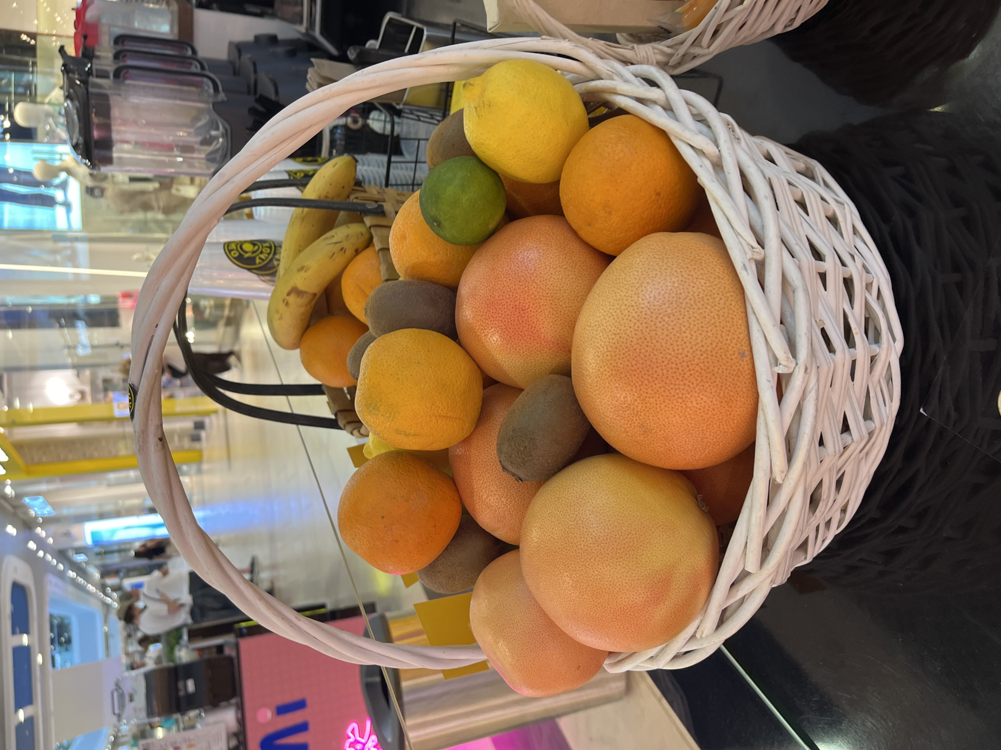 A close-up, eye-level, vertical shot shows a white woven basket filled with various fruits, predominantly grapefruits and oranges, with some kiwis, a lime, and a lemon also visible. The basket's handle, made of twisted white material, is prominent in the foreground, arching over the fruit. The fruits are arranged in a dense, layered fashion, filling the basket to the brim. The lighting is bright, casting subtle shadows on the fruits.
In the background, the scene transitions to a brightly lit indoor environment, resembling a shopping mall or a marketplace. Several tall, transparent blenders are visible to the right, suggesting a juice bar or a smoothie shop. To the left, a pink display with the word "Ni" in large blue letters is partially visible. In the mid-ground, blurred figures of people, dressed casually, are walking through the aisle. The overall impression of the background is that of a commercial setting with a clean, modern aesthetic.
This image was taken at the "Moy Island" shopping center in Sochi, Russia. The specific location within the shopping center appears to be an area dedicated to fresh juices or smoothies, given the prominent display of blenders and the basket of fruits. The time of day is likely daytime, indicated by the bright interior lighting.