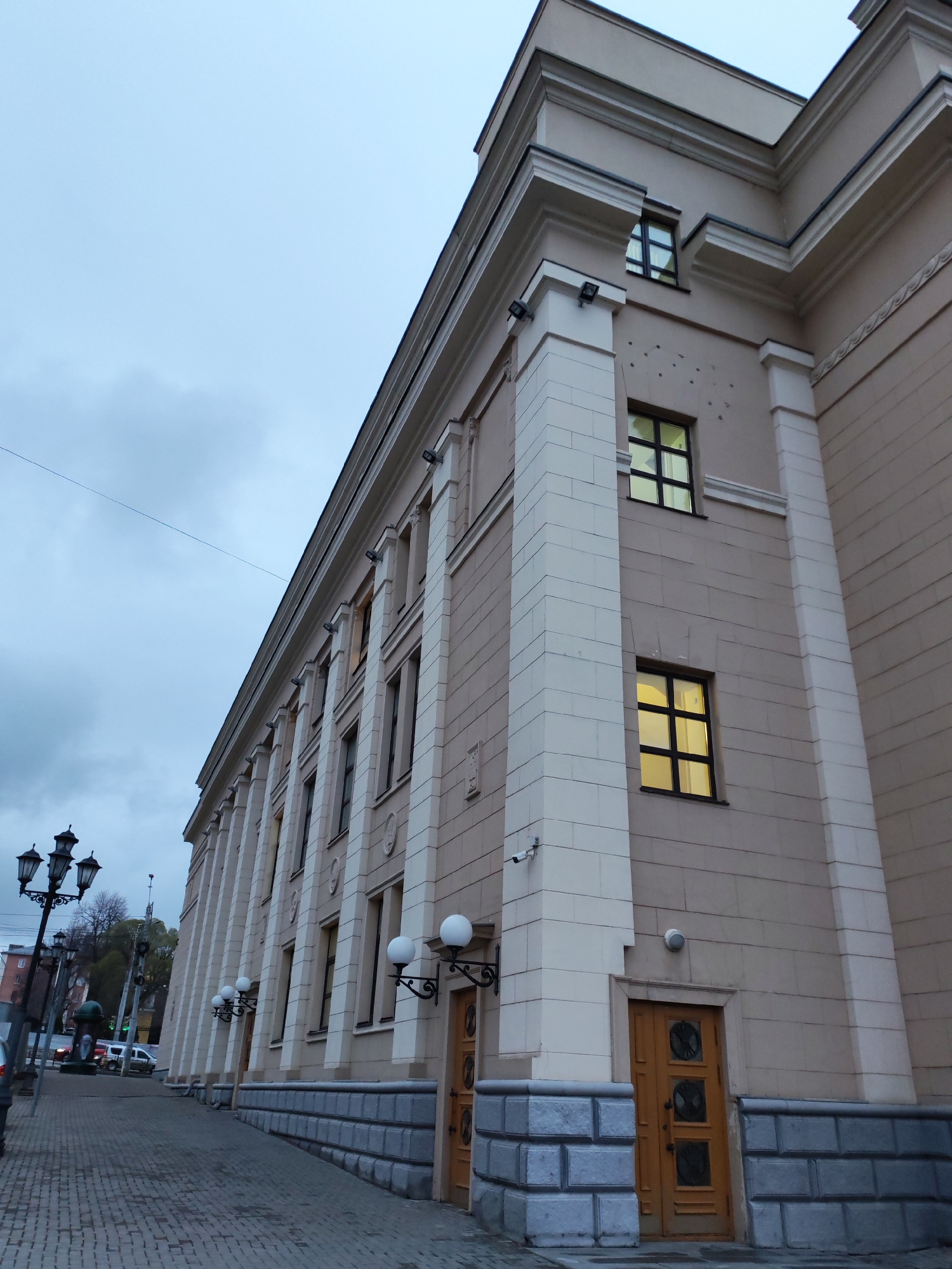 The image captures an exterior view of a building in Izhevsk, Russia, identified as the State Opera and Ballet Theatre of Udmurtia. The perspective is from the street level, looking up at an angle towards the facade of the building.
The building exhibits classical architectural elements, with a beige exterior complemented by white, column-like vertical sections that run along its length. Windows are visible at regular intervals, some with yellow light emanating from within. The building's base is lined with gray stone brickwork.
The street is paved with interlocking stone bricks, and there is a sidewalk adjacent to the building. A few vehicles are visible in the background. Along the street, there are antique-style street lamps with multiple globes.
The sky above is overcast.
There are no people directly visible in the image.