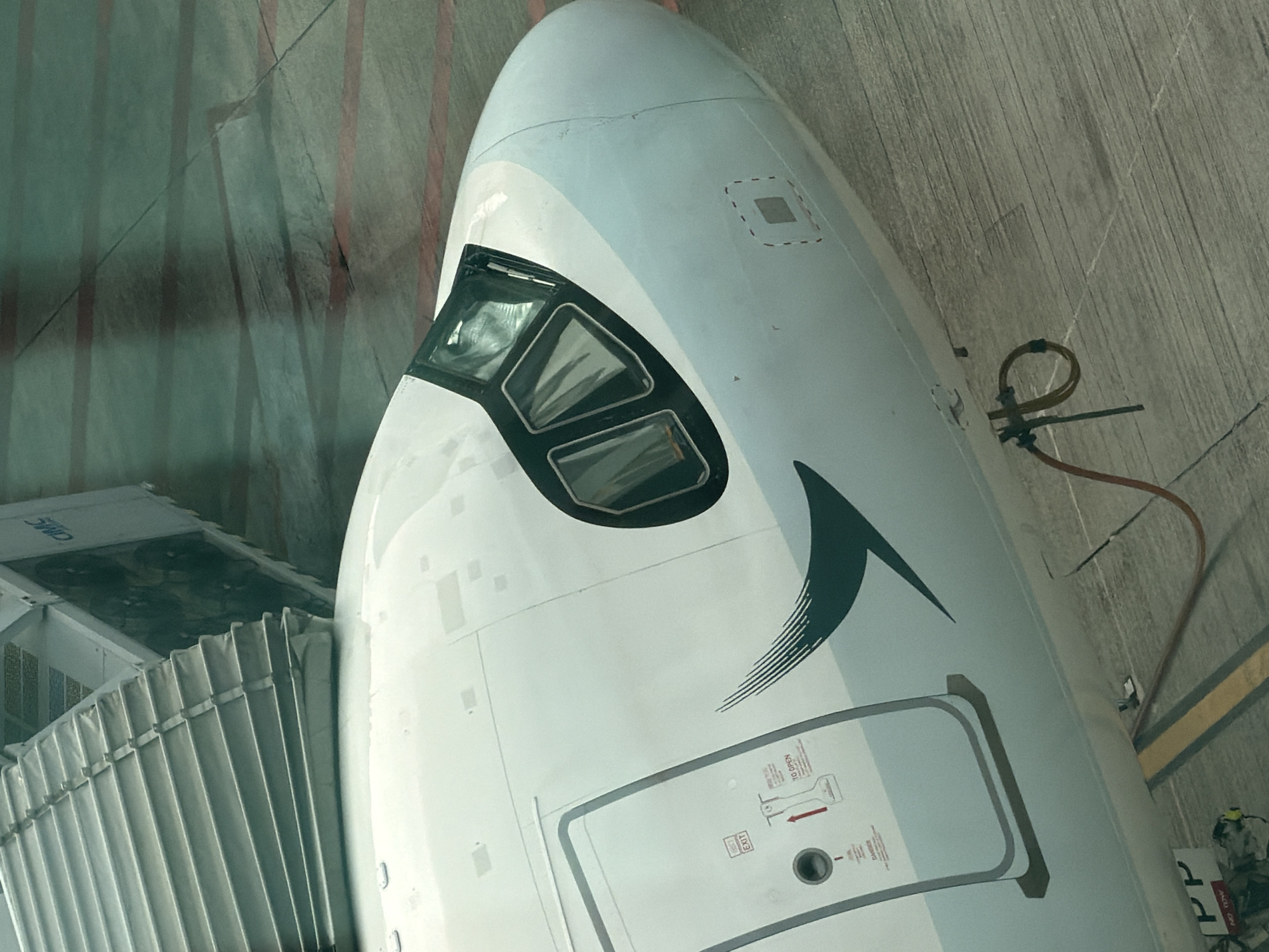 An airplane is parked at an airport gate. The front of the airplane is visible, showing the cockpit windows and part of the fuselage. A jet bridge is connected to the airplane. The ground is paved with concrete and has red lines painted on it, possibly indicating taxiways or parking spots. The overall scene suggests an airport environment. The airline livery is partially visible, featuring a green curved design on the fuselage. The text on the airplane's door includes "EXIT" and instructions for opening. The environment appears to be outdoors. Based on the airline's livery (Cathay Pacific, known for its green brushstroke logo) and the general setting, this image was likely taken at Hong Kong International Airport.
