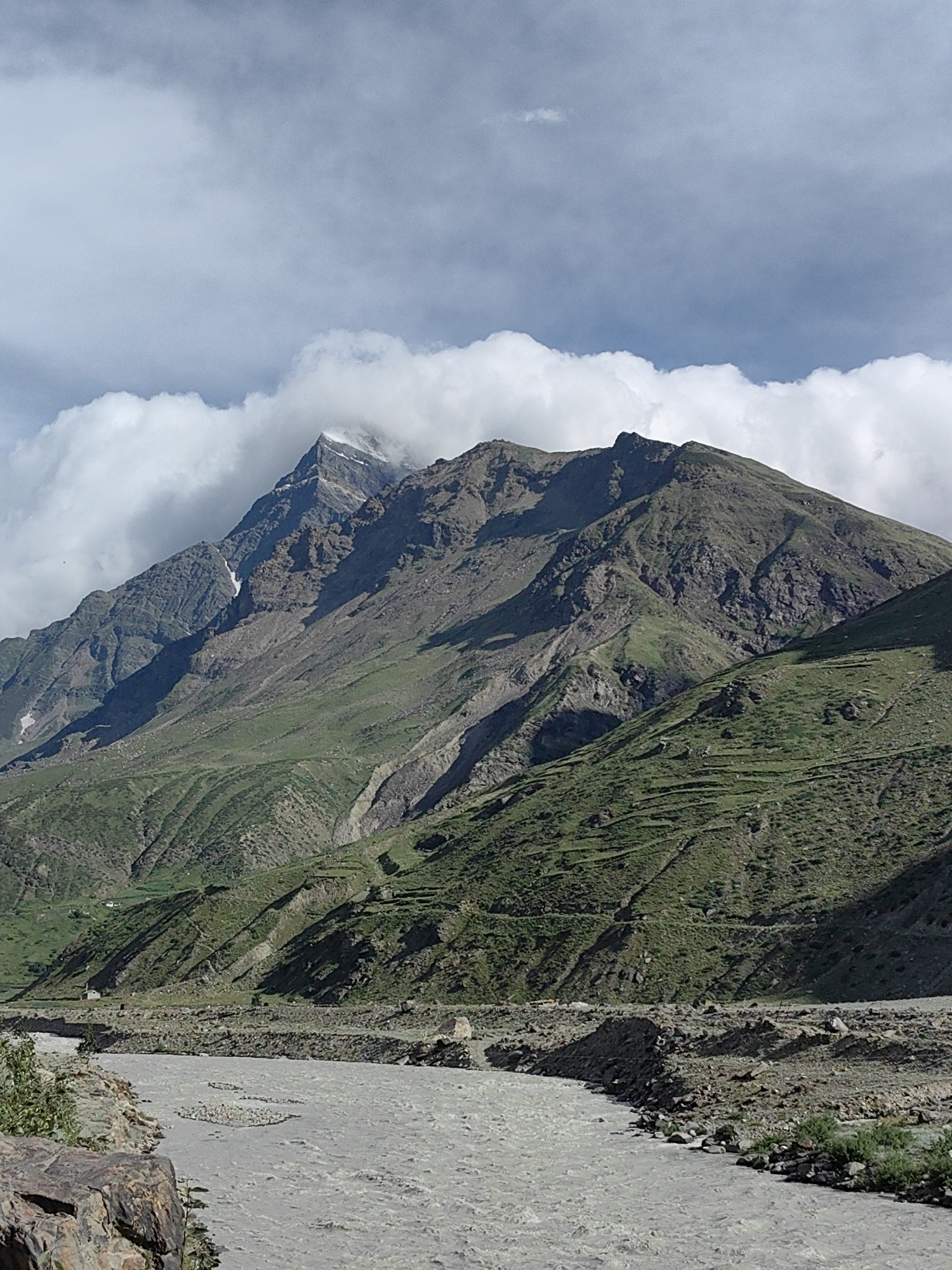 The image depicts a mountainous landscape near the village of Yamling, India. A wide, shallow river with murky, silt-laden water flows through the foreground. The riverbanks are strewn with rocks and gravel. In the midground, the steep slopes of a mountain rise dramatically. These slopes are covered in green vegetation, with evidence of terraced farming or natural formations creating distinct lines. The mountain itself is rugged and rocky, with some patches of snow or ice visible near its peak. The upper part of the mountain is partially obscured by large, white cumulus clouds that are drifting across a pale blue sky. The overall scene is one of natural grandeur and rugged terrain typical of a mountainous region. There are no people or identifiable structures clearly visible in this specific frame, beyond what appears to be some very small, distant buildings or settlements along the mountain slopes.
