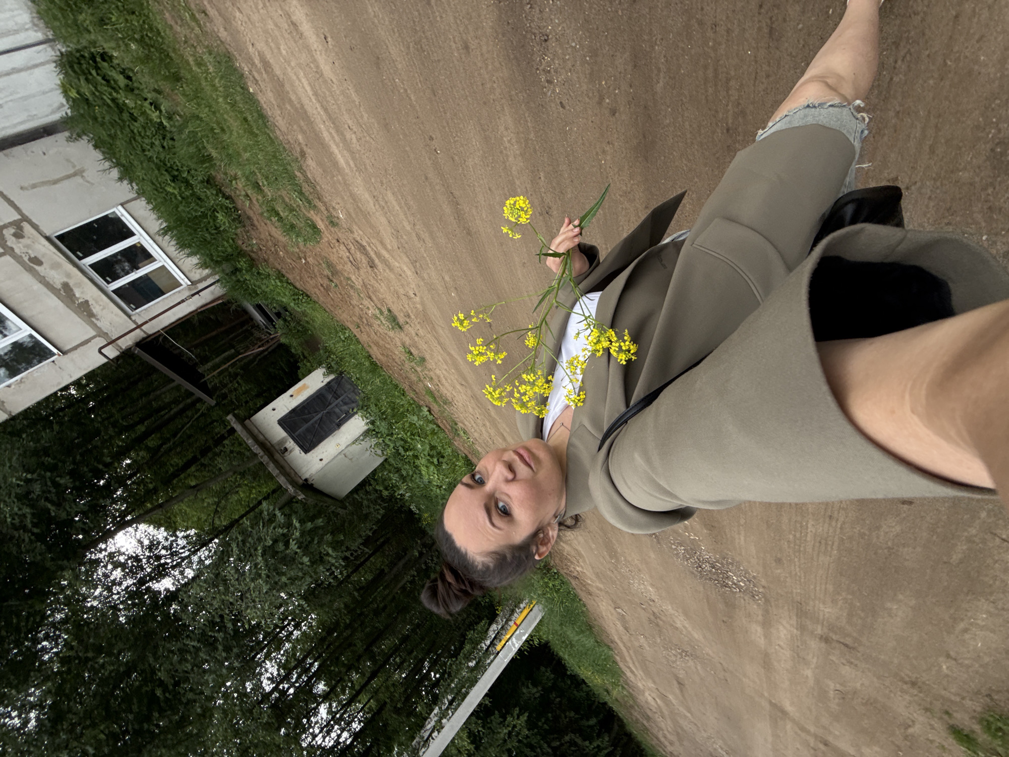 The image is a full-body selfie of a woman taken outdoors in a rural or semi-rural setting, likely in Chaikovsky, Russia. The photo is angled, creating a Dutch tilt effect.
The main subject is a woman with dark hair tied in a bun and dark eyes, looking directly at the camera with a neutral expression. She is wearing a light-colored top under an olive green or khaki blazer/jacket. In her right hand, she holds a sprig of bright yellow wildflowers, possibly rapeseed. Part of her left leg is visible, appearing to be clad in distressed denim shorts or jeans under the blazer.
The background features a wide, unpaved dirt road that occupies a significant portion of the scene. To the left, there is a light-colored, possibly concrete or stucco, building with several rectangular windows. Further back and to the left, a dense area of dark green trees suggests a wooded or forested area. Another smaller, squarish structure with a dark top is visible amongst the trees. Green grass and foliage line the edges of the dirt road and surround the buildings. A metallic pole or beam is partially visible in the lower-left quadrant of the image.
The overall lighting suggests it is daytime, possibly overcast, creating a soft, diffused light. There is no visible text in the image. The setting appears calm and natural, with elements of simple architecture.