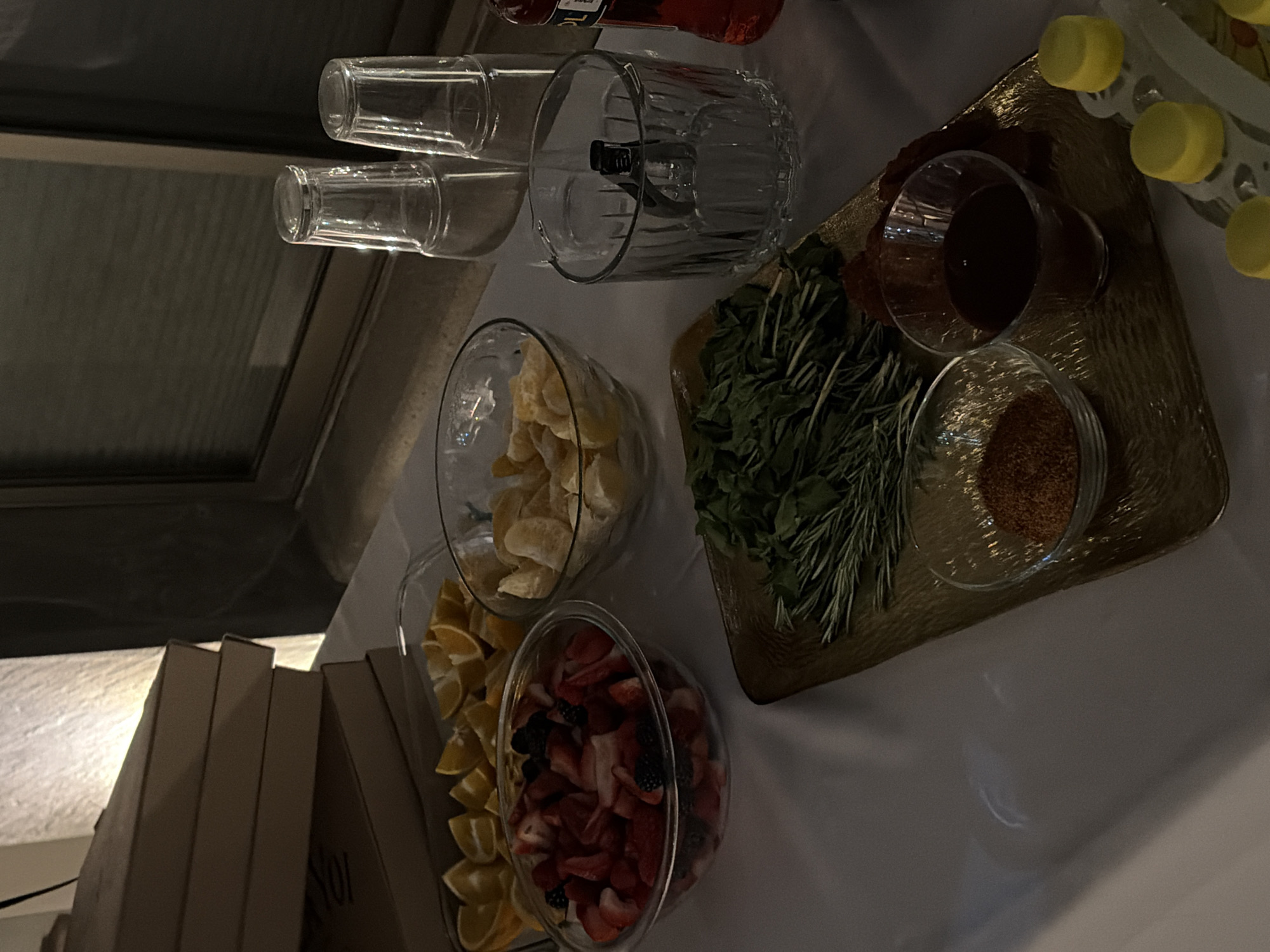 The image depicts a dimly lit event setup featuring food and beverage ingredients on a white tablecloth. In the upper left, a stack of four light brown cardboard boxes, possibly pizza boxes, rests beside a textured, illuminated wall. To the right, a dark window frame with horizontal blinds is visible.
On the table, an array of ingredients is arranged in clear glass bowls: a rectangular dish contains sliced orange segments, a round bowl holds diced strawberries and blackberries, and another round bowl contains peeled orange or tangerine segments.
Towards the right, two inverted, clear drinking glasses are stacked, and a single ribbed glass, containing what appears to be ice tongs, stands below them. A partially visible red-orange bottle is positioned on the far right.
A textured gold-colored tray in the foreground holds fresh herbs, specifically bunches of rosemary and broader-leafed herbs like mint or basil. On this tray, two small clear glass bowls are present: one filled with a reddish-brown spice blend (possibly chili powder) and another containing a dark brown liquid (likely a sauce or syrup). Dark, irregular shapes, possibly dried chilies or cured meats, are visible behind these bowls on the tray. In the bottom right corner, a white dispenser with multiple bright yellow capped items, potentially small bottles or condiments, is partially visible.
No individuals are present in the scene, and no specific actions are depicted beyond the arrangement of items. The setting appears to be an indoor or semi-outdoor space, possibly a hospitality venue, consistent with event preparation. The image does not contain any specific visual cues, such as landmarks or identifying signage, to pinpoint an exact location within the city of Puebla, Mexico.