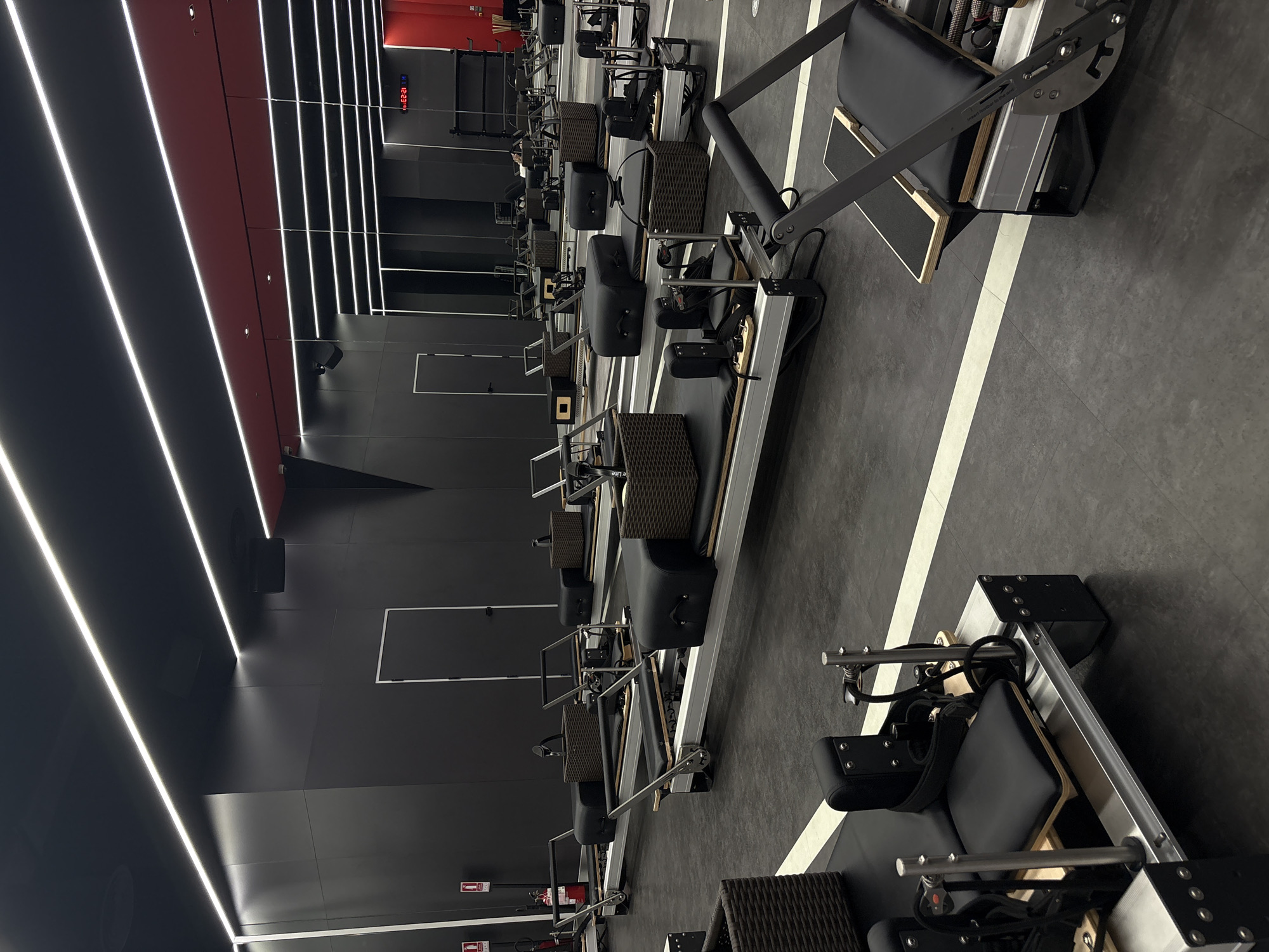 The image shows a row of black pilates reformers in a modern, dimly lit fitness studio. The reformers are lined up parallel to each other on a dark grey floor with white lines. The studio features sleek black walls and a ceiling with bright white LED strip lighting, creating a contemporary and high-tech atmosphere.
There are no people visible in the image. The pilates reformers are the primary objects, each equipped with a sliding carriage, footbar, springs, and straps, indicating they are ready for use. Some reformers have woven baskets on the side. The lighting suggests it could be evening or a space with controlled lighting, regardless of the time of day outside.
The location context provided is Nong Pa Khrang, Thailand, but there are no specific visual cues in the image to confirm this location. No weather conditions are discernible. The overall mood conveyed is one of order, cleanliness, and preparedness for a workout.
There is some visible text: a digital clock displaying "11:22:28" is mounted on the wall in the background. There are also some small red signs with white symbols that appear to be fire exit signs, common in public spaces. One of the pilates reformers has text that is partially visible and reads "insert buffer to mat".