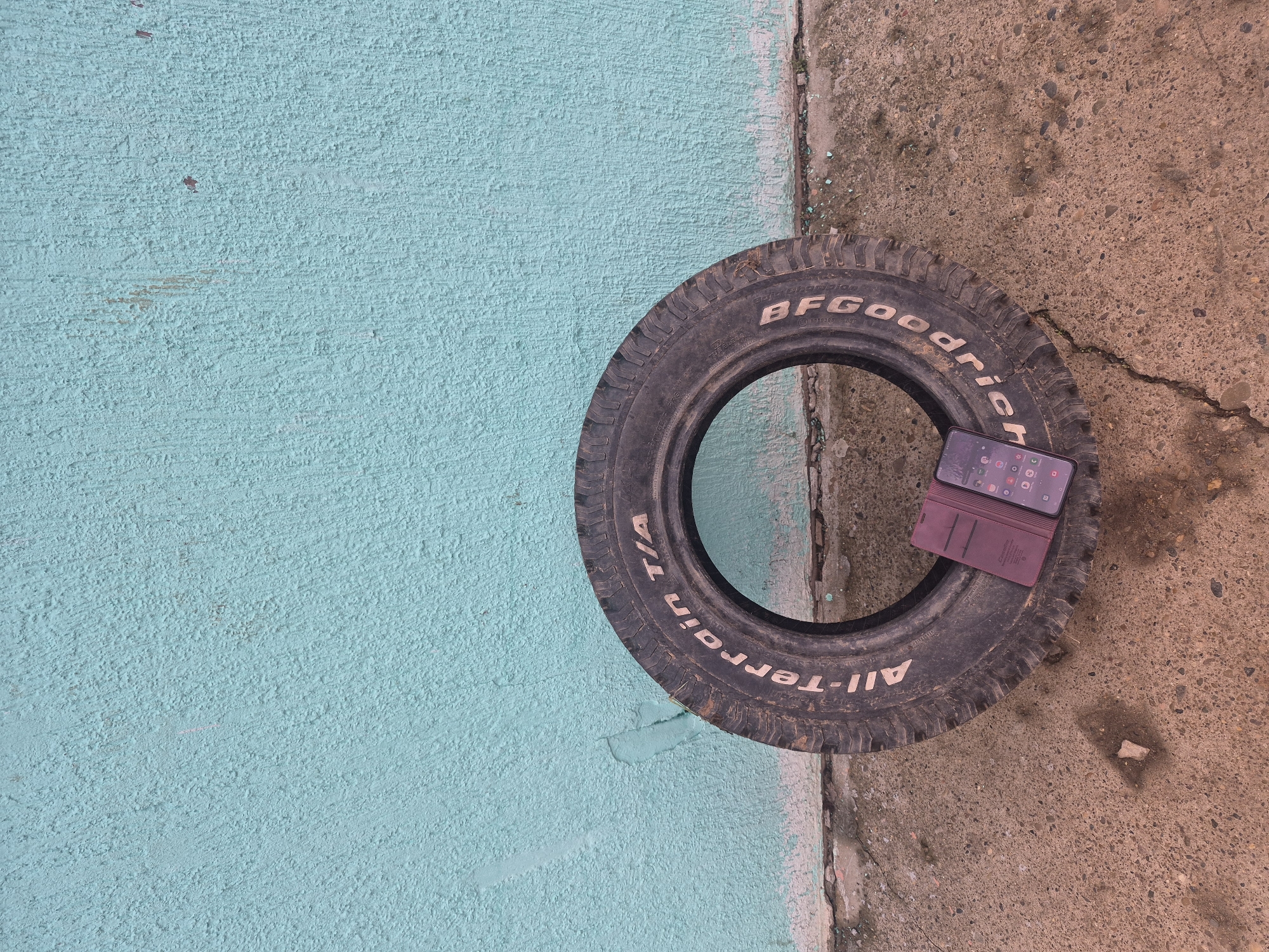 The image shows a discarded BFGoodrich All-Terrain T/A tire lying on a surface that transitions from a light blue textured wall on the left to a worn, mottled concrete surface on the right. A smartphone with a maroon-colored case is situated inside the tire's inner circumference. The location appears to be outdoors in an undefined urban setting, Unknown, Unknown. No people are visible in the image. The tire is positioned at an oblique angle across the boundary between the blue wall and the concrete.