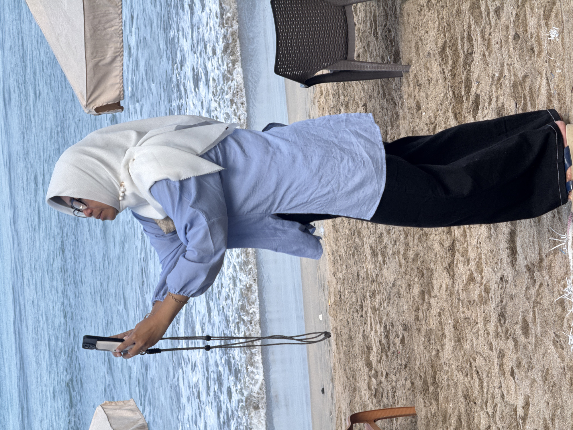 A woman stands on a sandy beach in Kuta, Indonesia, taking a selfie with her phone. She is wearing a light blue long-sleeved shirt, black pants, and a white hijab. A cord from her phone dangles down her front. The ocean with small waves is visible behind her. To her right, a dark plastic chair sits on the sand. To her left, the edge of a cream-colored beach umbrella is visible. The sand appears damp and textured, with some debris scattered on it.
