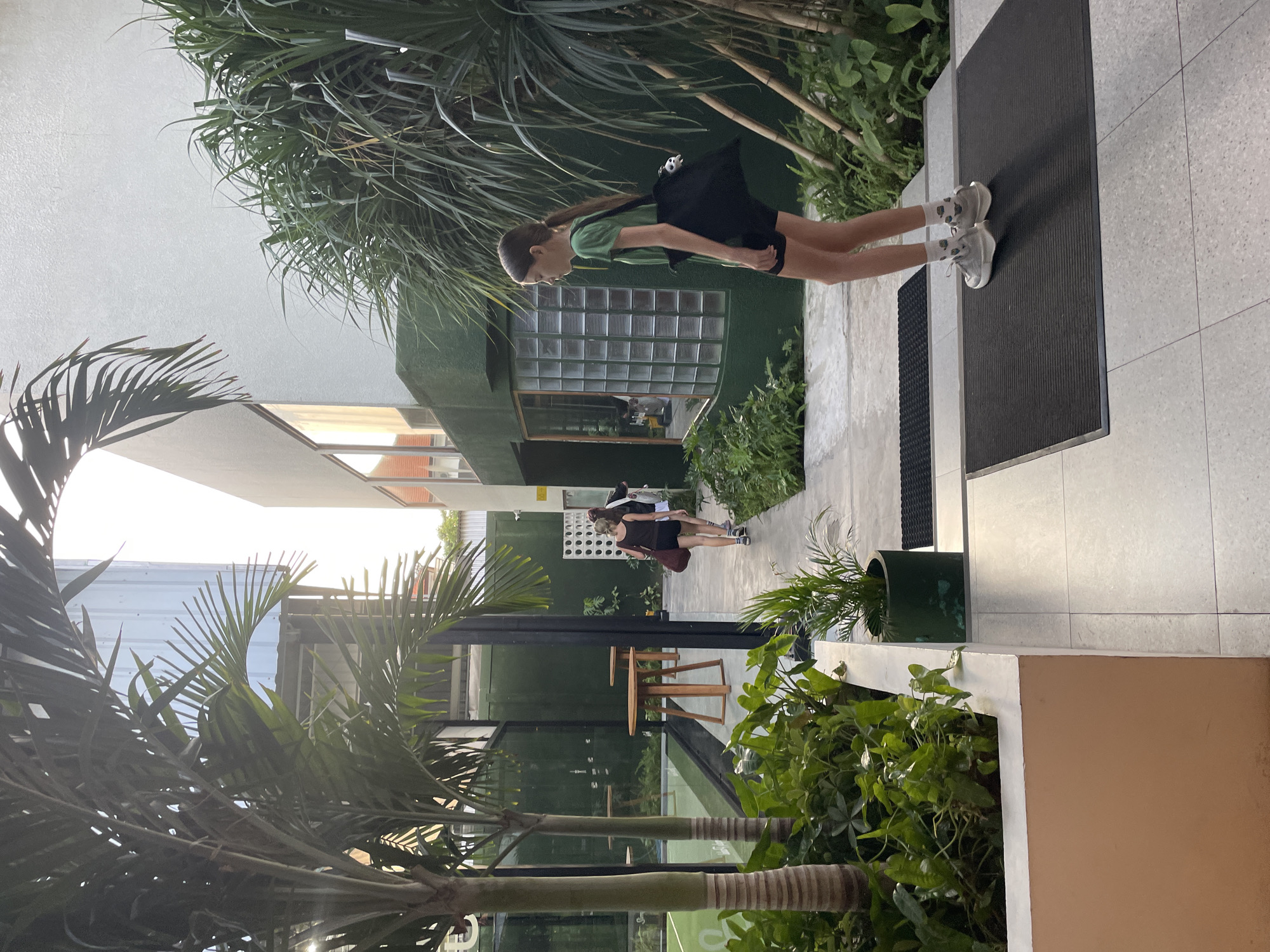 A vertical shot presents a sunlit scene in what appears to be Denpasar, Indonesia. A girl with long brown hair, wearing a green t-shirt, black shorts, white socks, and white tennis shoes, and carrying a black bag, walks on a tiled platform, stepping onto a rectangular doormat. Further down a shaded path, two women are seen walking away from the camera. The foreground includes tropical foliage: palm trees and various green plants, some potted in cylindrical green planters. Beyond the path, a building with a partially visible tennis court can be seen. Light bleeds into the image through the palm fronds, adding contrast. The overall environment suggests an outdoor, possibly urban, setting.