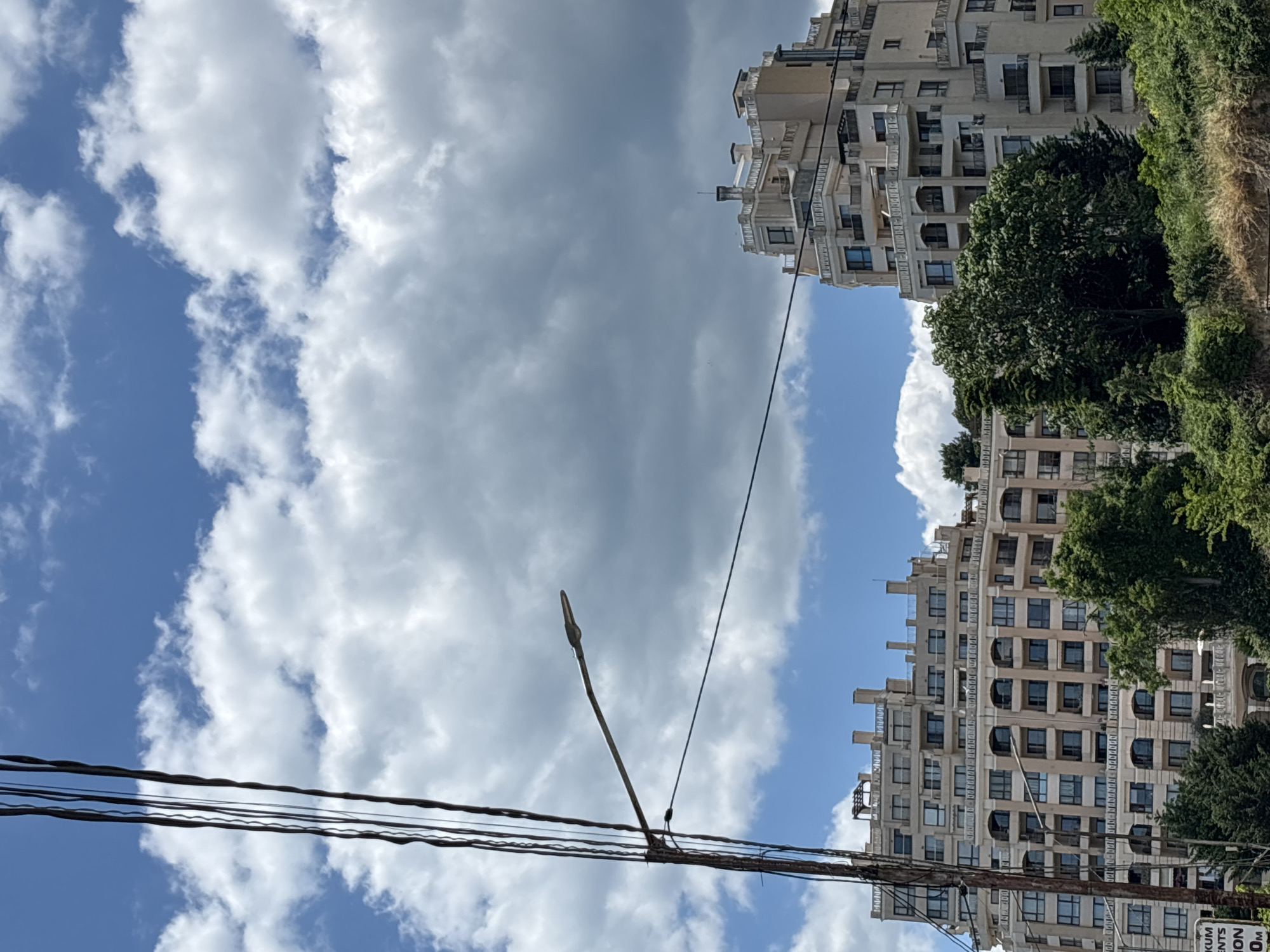 The image shows a view of multi-story buildings and trees against a cloudy blue sky. In the foreground, a utility pole with multiple wires and a street lamp is visible on the left. To the right of the pole, a large, light-colored apartment building with many balconies and windows dominates the mid-ground. Behind it and to the left, another similar building is partially visible. Lush green trees are in front of and between the buildings. The scene appears to be taken during daylight. The architecture suggests a modern residential area, possibly in a resort town. Given the characteristic architectural style of buildings like these, this is likely a view of the Grand Mall area in Varna, Bulgaria, specifically looking towards the residential complexes that surround it.