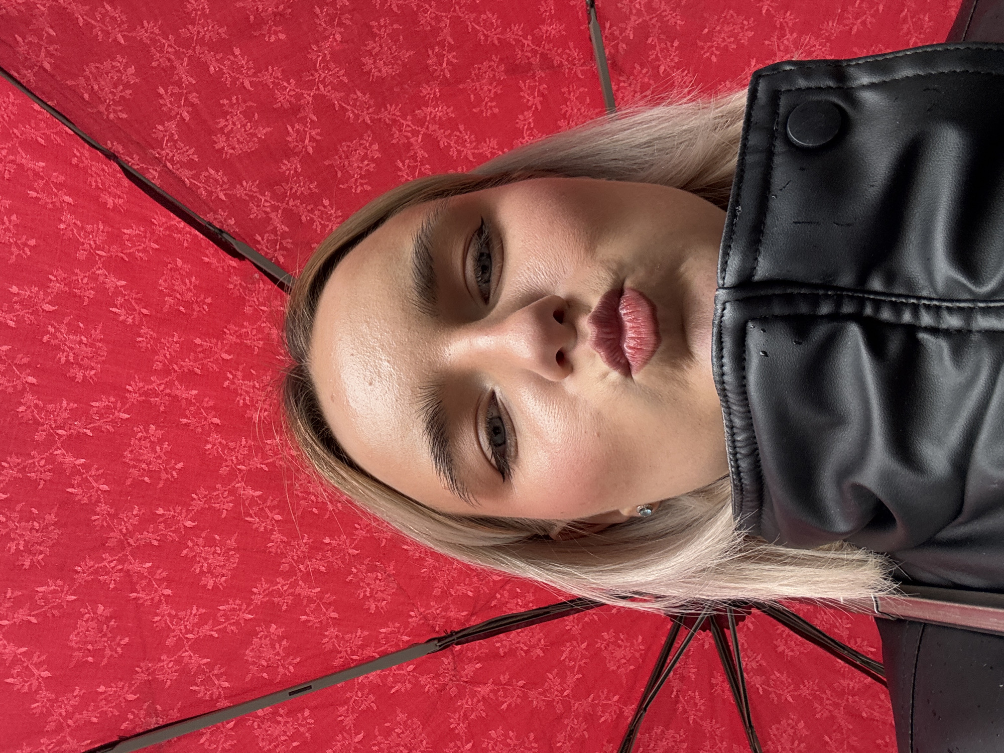 A woman is positioned under a red umbrella with a floral pattern. She has blonde hair, light skin, and is wearing a black leather jacket with a button on the lapel. The background is the inside of the umbrella, which is red with a recurring floral pattern. The location cannot be determined from the image.
