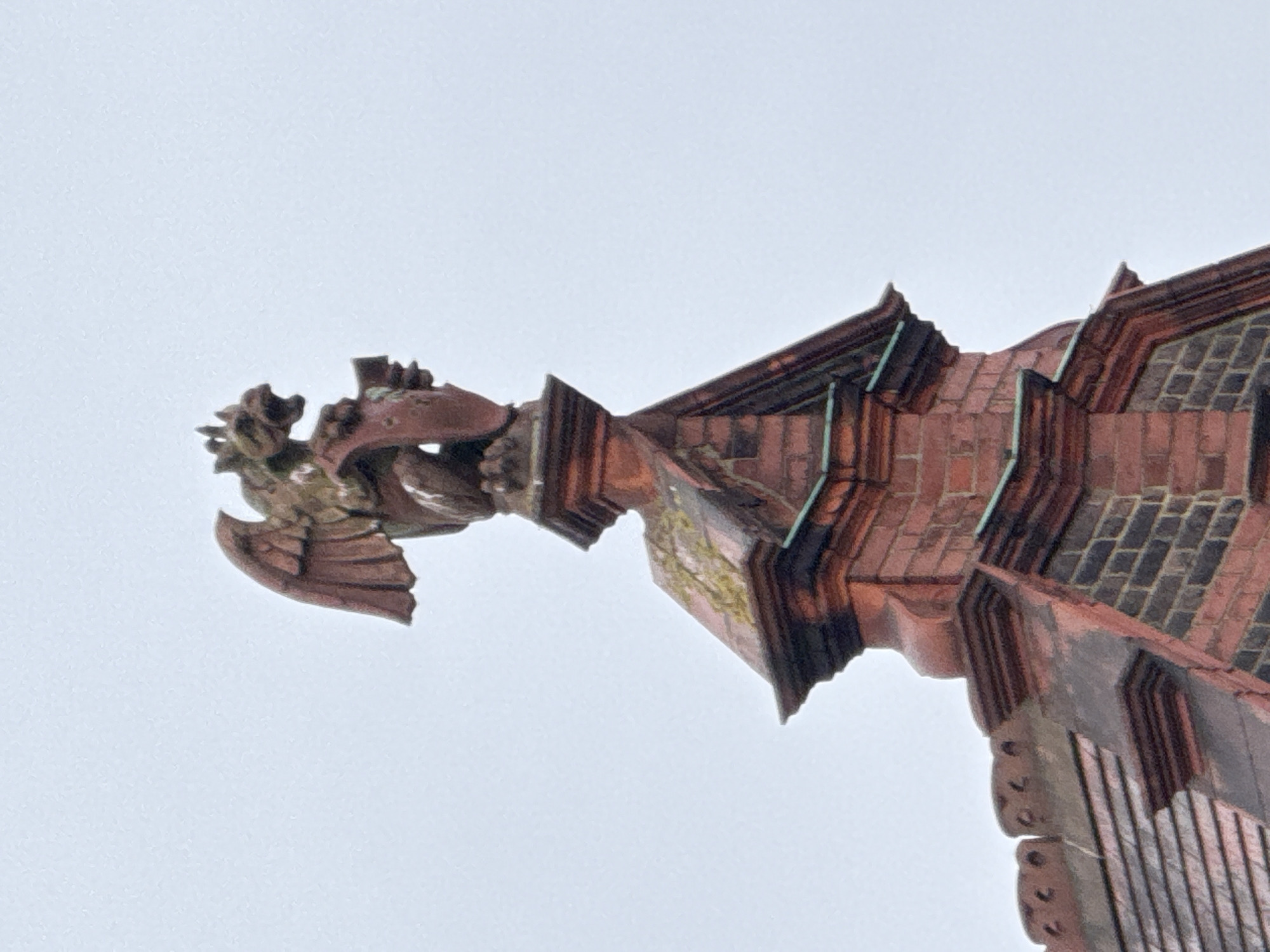 The image shows a gargoyle protruding from a red brick building. The gargoyle has wings and an ornate head. The building's architecture suggests a historical or gothic style with detailed brickwork. The sky is overcast and grey. It is possible this image was taken in London, United Kingdom due to the style of the architecture, but further details would be needed to confirm this.