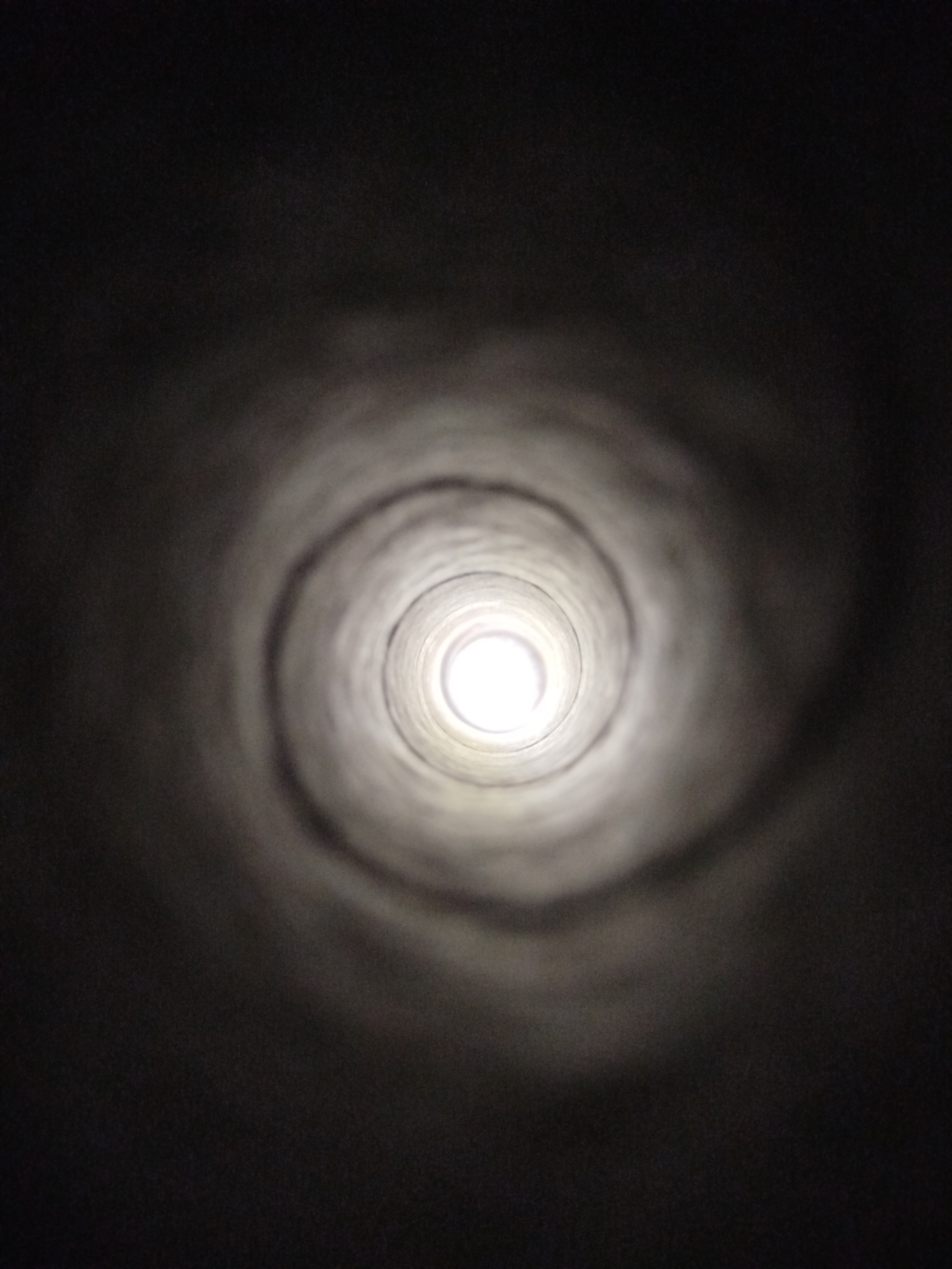 The image is a low-light photograph taken from inside a cylindrical tunnel or pipe, looking towards a bright light source at the far end. The tunnel walls are textured and appear to be made of concrete or a similar material, with swirling patterns visible due to the lighting. The overall mood of the image is dark and somewhat mysterious, with the bright light at the end creating a sense of depth and focus. The specific location in the city of Kremenchuk, Ukraine, cannot be determined from the image alone, as it lacks any distinguishing landmarks or contextual clues. The photograph is a close-up, abstract view of a man-made structure, with no discernible people, animals, or specific objects other than the tunnel itself. The composition is symmetrical, with the light source at the center and the tunnel walls radiating outwards. The image is likely intended to evoke a sense of perspective or a journey into the unknown.