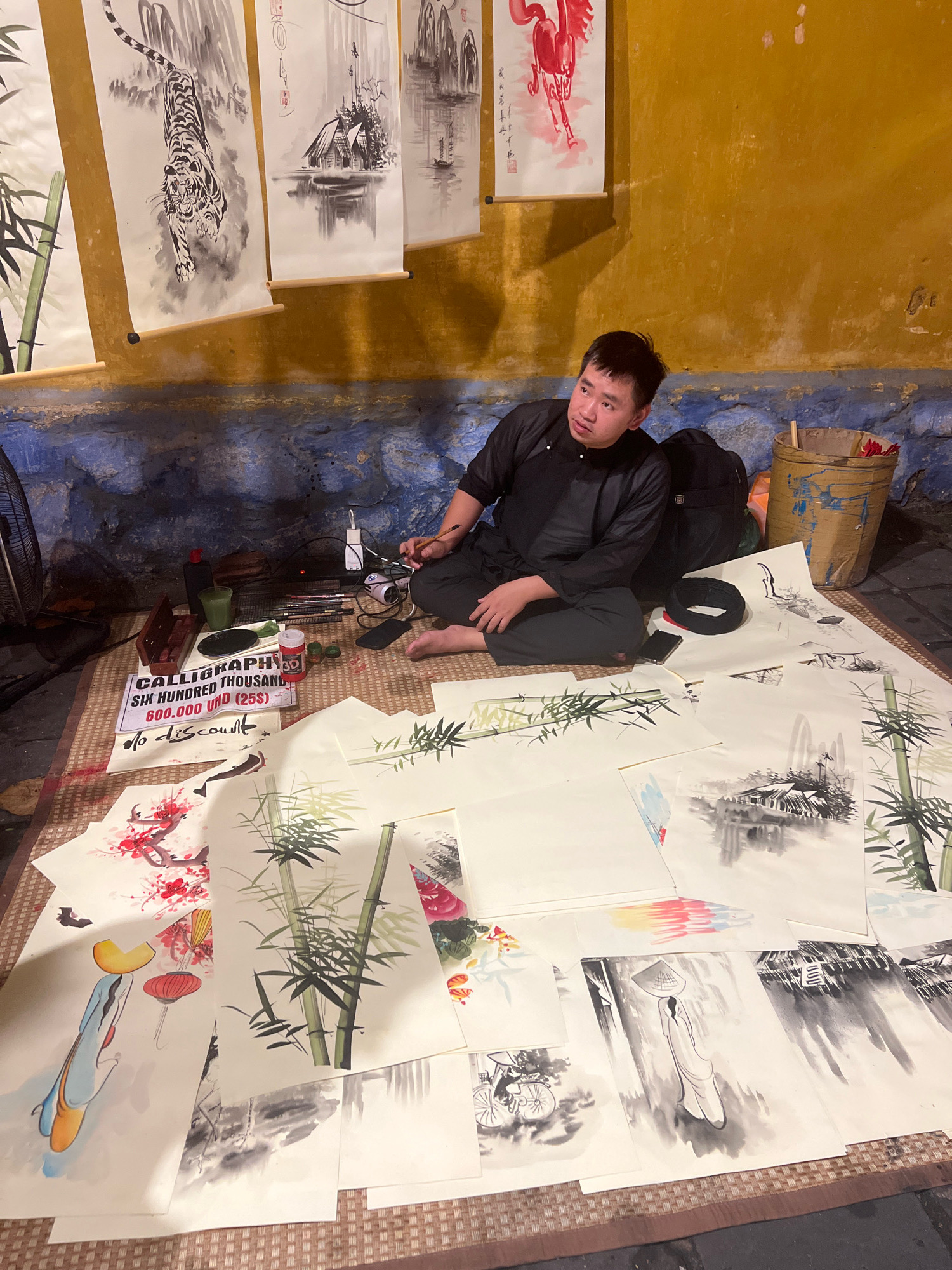 The image depicts an outdoor scene in Hoi An, Vietnam, where a young man is seated on the ground surrounded by various ink paintings. He is wearing traditional black clothing. The paintings, displayed on scrolls and as individual sheets, feature subjects such as tigers, bamboo, landscapes, and figures. Among the objects on the ground are brushes, ink bottles, a cup, and a sign indicating "CALLIGRAPHY SIX HUNDRED THOUSAND 600.000 VND (25$)" with "no discount". The background shows a textured, yellow-ochre wall with a blue, rough-textured lower section. A basket and a backpack are also visible. The overall setting suggests a vendor or artist preparing to sell or display their artwork in a public space.