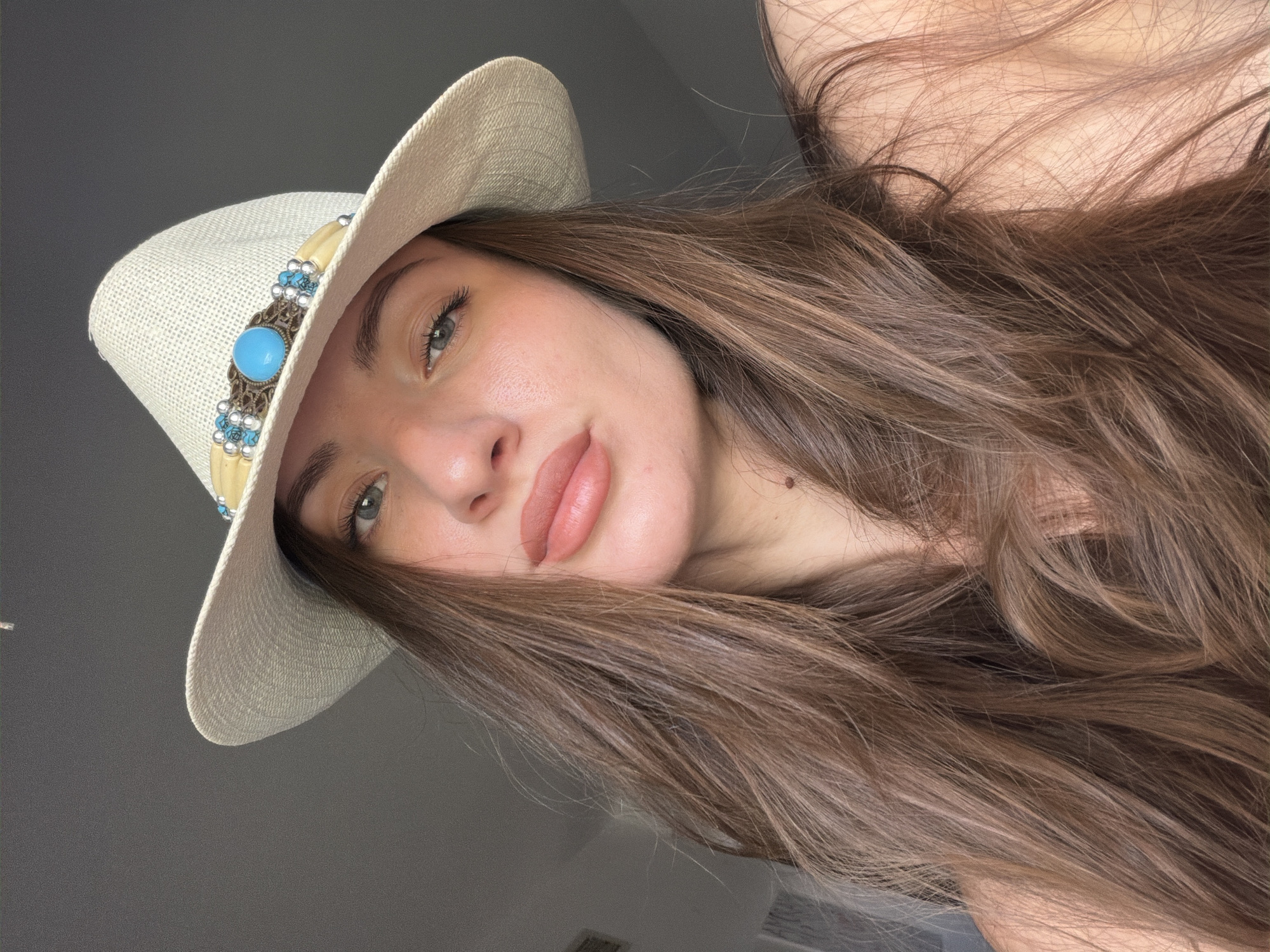 A close-up, selfie-style, headshot of a young woman wearing a straw fedora hat adorned with a turquoise stone and beaded detailing. Her long, brown hair is loose and cascades around her shoulders. The woman has fair skin, blue-green eyes, and full lips. She is looking directly at the camera with a neutral expression. The background is a plain, grey wall. The image does not contain any information that places it in Sharm El Sheikh, Egypt.
