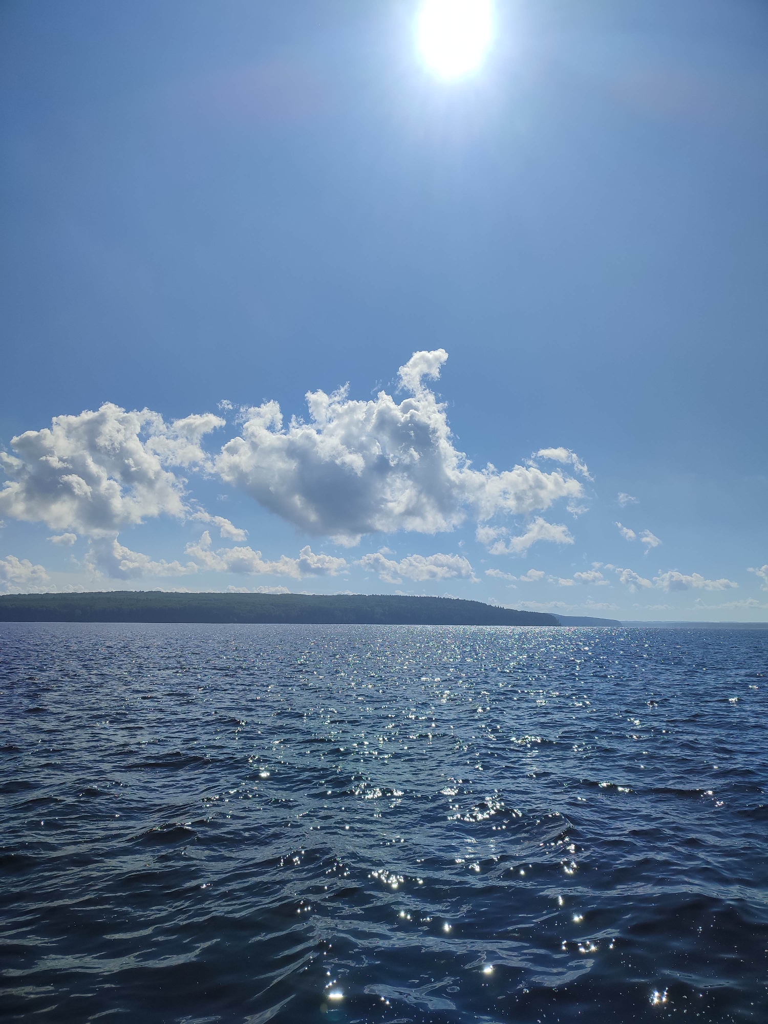 A bright, sun-drenched scene depicts a vast expanse of deep blue water rippling under a clear sky. The sun, a radiant white orb, shines intensely from the upper right quadrant of the image, casting a shimmering effect across the water's surface. A line of dark green, tree-covered land forms a distant horizon across the center of the frame. Fluffy white clouds are scattered across the bright blue sky, with a larger cluster of cumulus clouds forming a prominent shape in the upper left. The water appears calm, with gentle waves creating a textured, sparkling appearance due to the sunlight. No people or man-made objects are visible in the frame. The overall impression is one of a serene and natural outdoor environment.