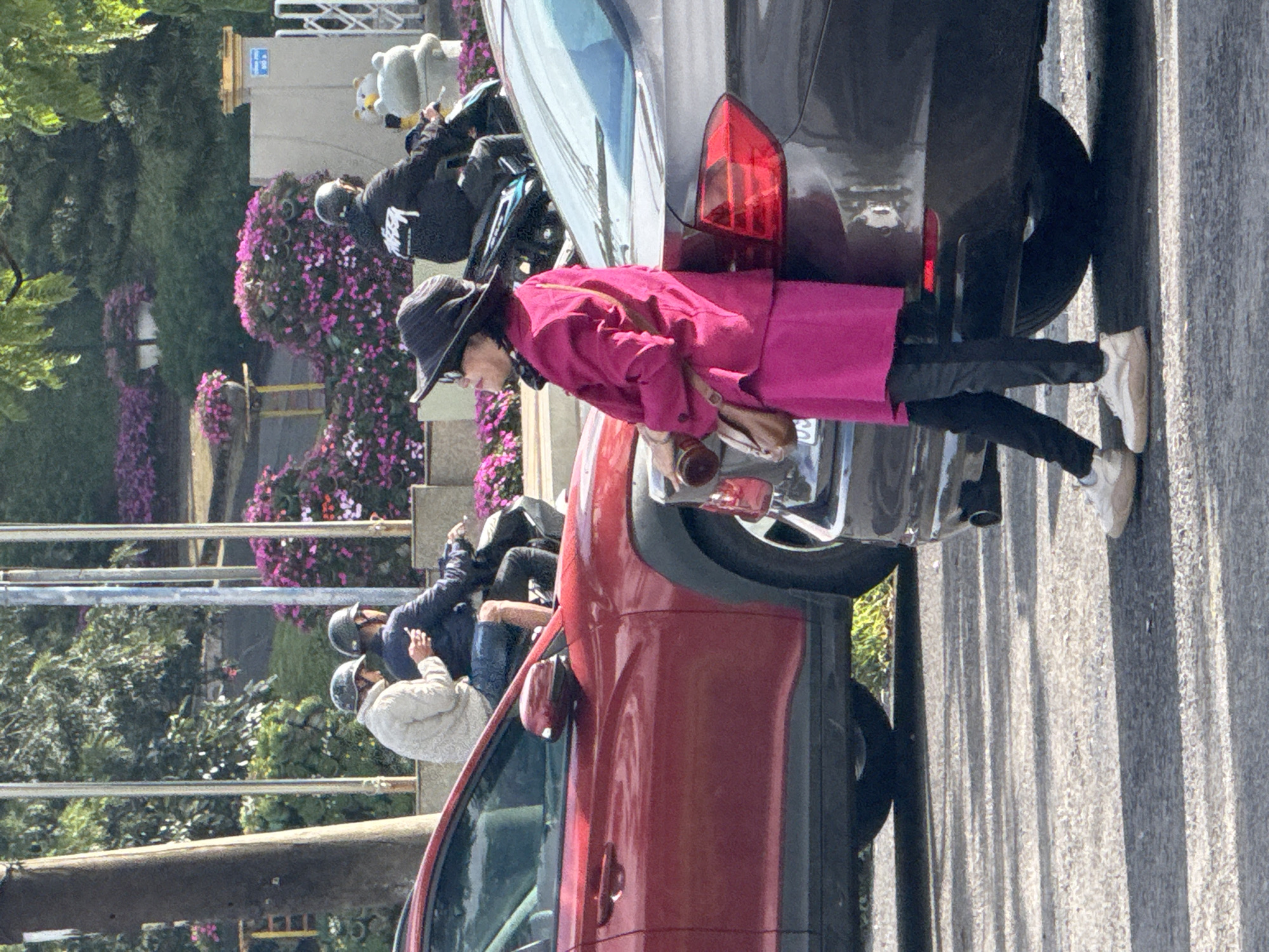 In Phường Xuân Hương - Đà Lạt, Vietnam, on a paved road, a woman in a pink blazer, black pants, and a black wide-brimmed hat stands near a dark gray car, holding a brown cylindrical object and a purse. She is facing left. Behind her, a person on a dark motorcycle wears a black hoodie with white lettering. To the left, two individuals on a motorcycle, both wearing helmets, are seated. One wears a light-colored jacket and holds a white object, possibly a phone. The background features lush green trees, vibrant pink flowers in planters, and a light-colored building with blue signage. The scene is illuminated by sunlight, casting shadows on the road.