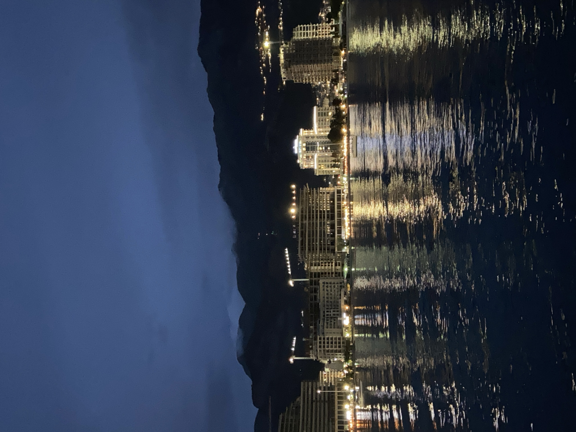 The image depicts a nighttime scene of the Budva skyline in Montenegro, reflected in the water. The city is situated at the base of a dark, mountainous landscape. Building lights illuminate the city, and their reflections create streaks of light in the water. The sky is a dark blue. There is no people in the image.