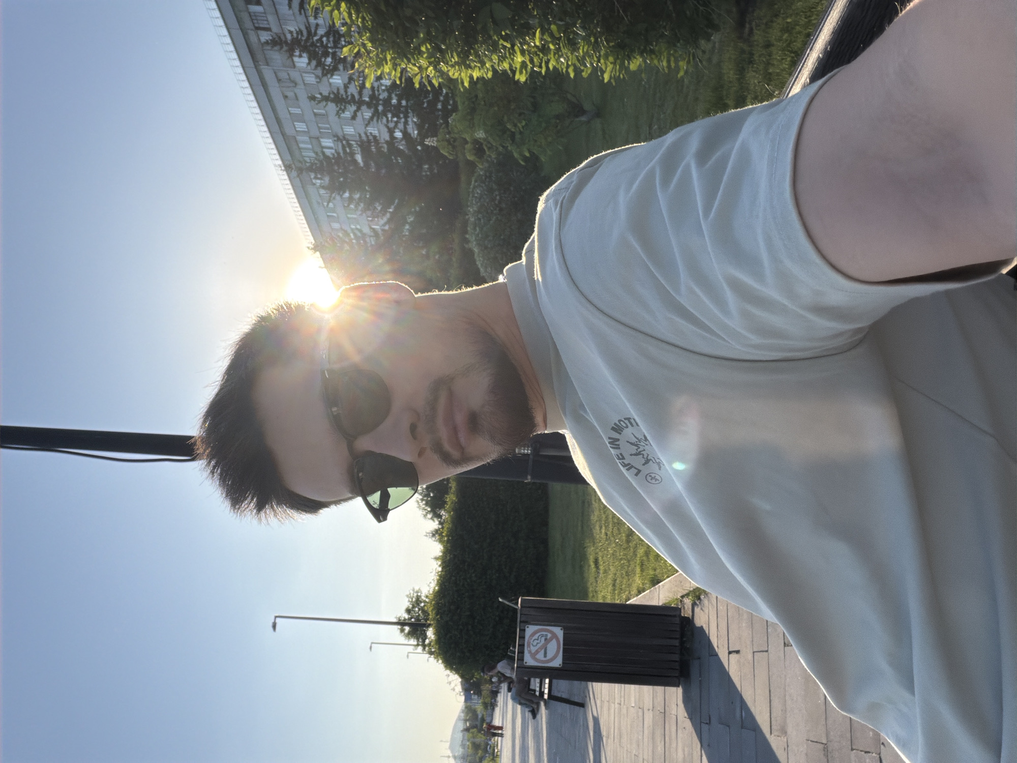 An outdoor selfie captures a male subject in a public urban setting during golden hour, likely sunset or sunrise, with the sun prominently visible behind his head, causing lens flare and backlighting. The image is rotated 90 degrees clockwise.
The subject is a man with short dark hair and a beard, wearing dark sunglasses and a light-colored t-shirt. He is positioned in the foreground, looking directly at the camera. A partially visible black graphic on his t-shirt sleeve appears to include the text "LIFE IN MOTTION" and some abstract shapes.
The background shows a paved walkway or promenade with lampposts. To the subject's right, a dark trash bin with a clear "No Smoking" symbol is visible. Further back, a wooden bench can be seen, with a person sitting on it. Lush green trees and bushes line the area. In the far distance on the left, a tall building with many windows is partially obscured by foliage. Faintly in the very distant background, a dome-like structure is discernible, possibly a landmark.
The sky is clear and bright blue, illuminated by the low sun. The scene suggests a park or waterfront promenade in an urban environment, consistent with the provided location context of Krasnoyarsk, Russia.